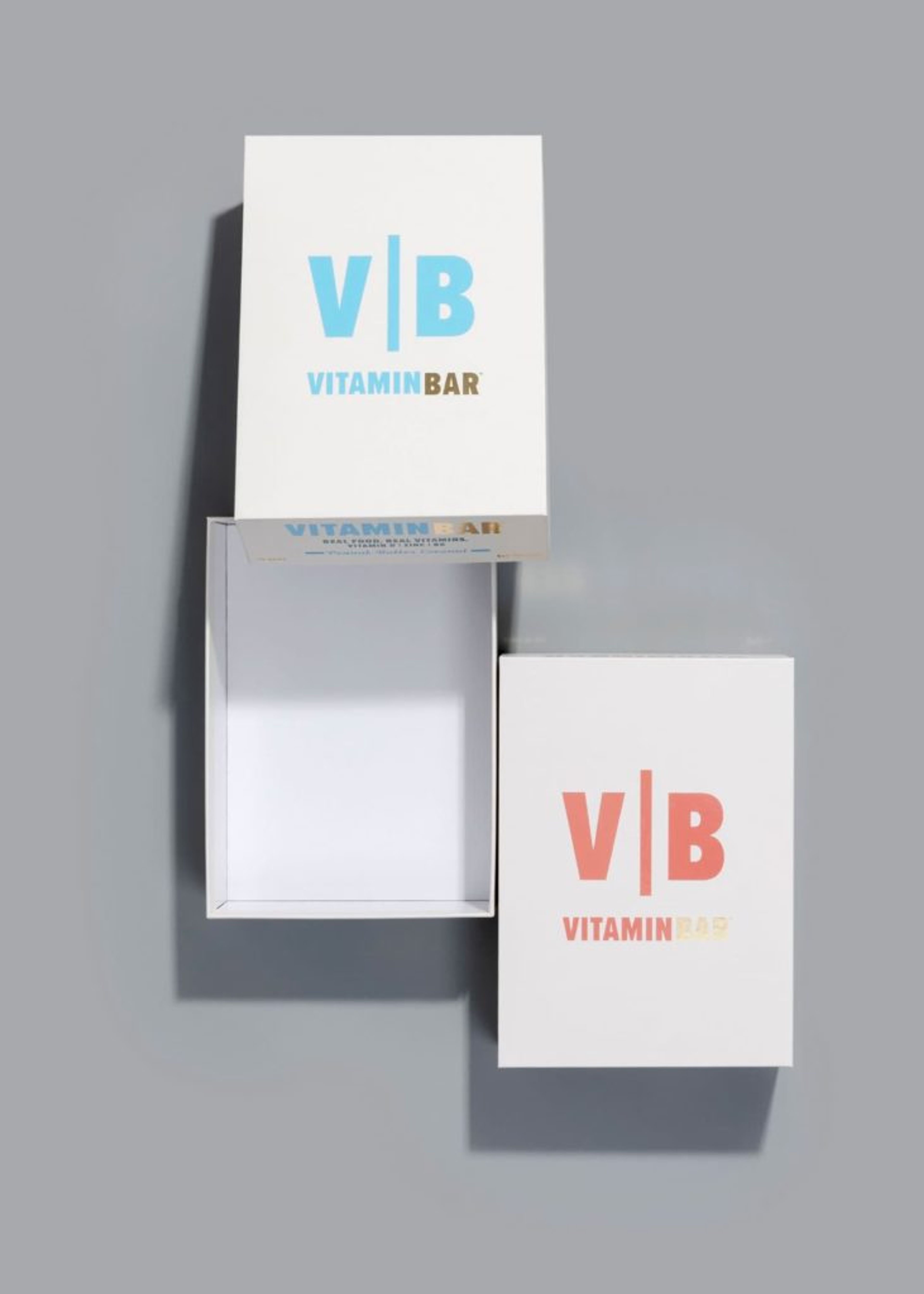 Top-down view of open and closed white Vitamin Bar boxes with blue and pink logos on a grey surface.