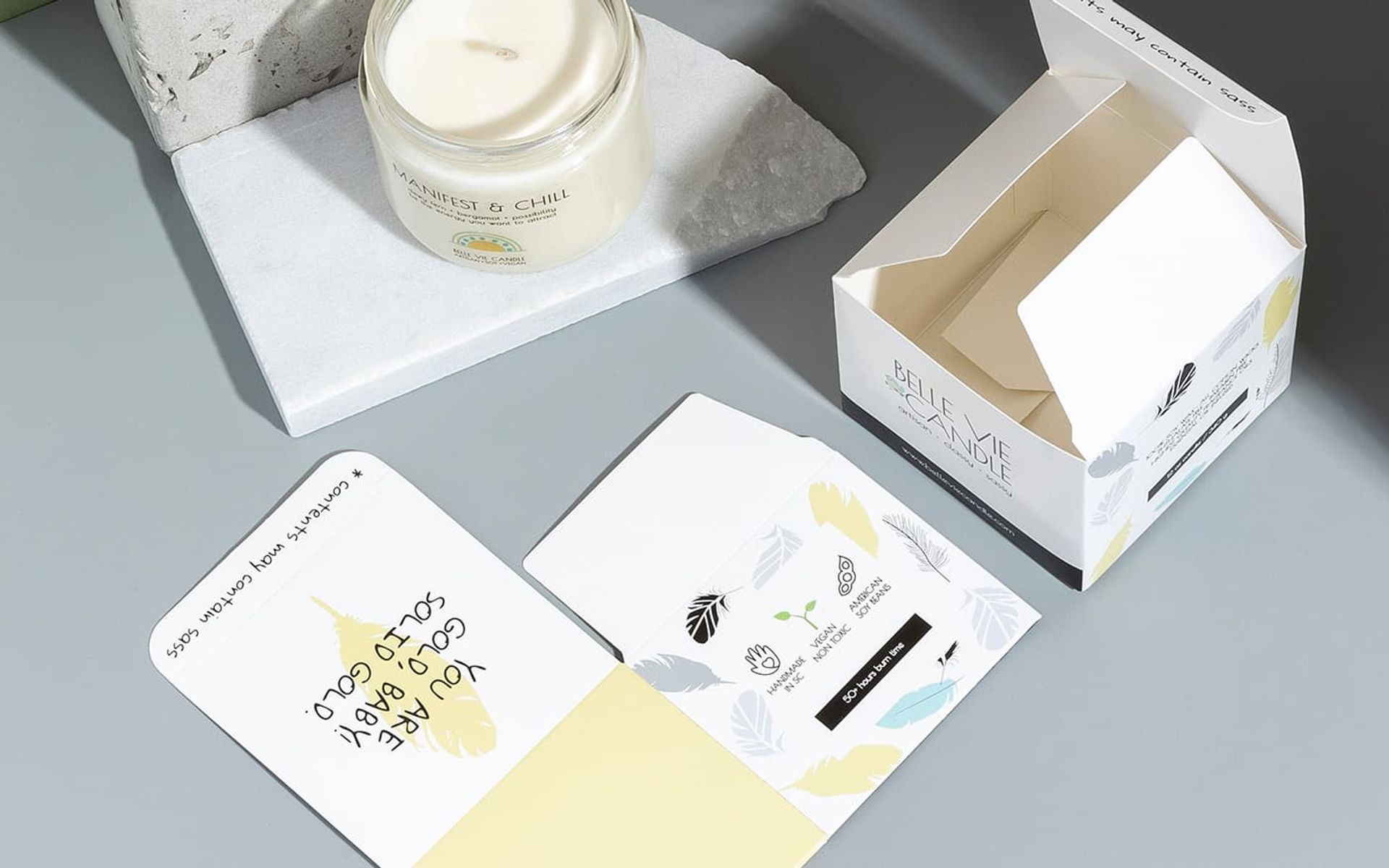 Top-down view of a lit Manifest & Chill candle next to an open Belle Vie box revealing hidden sassy interior text.