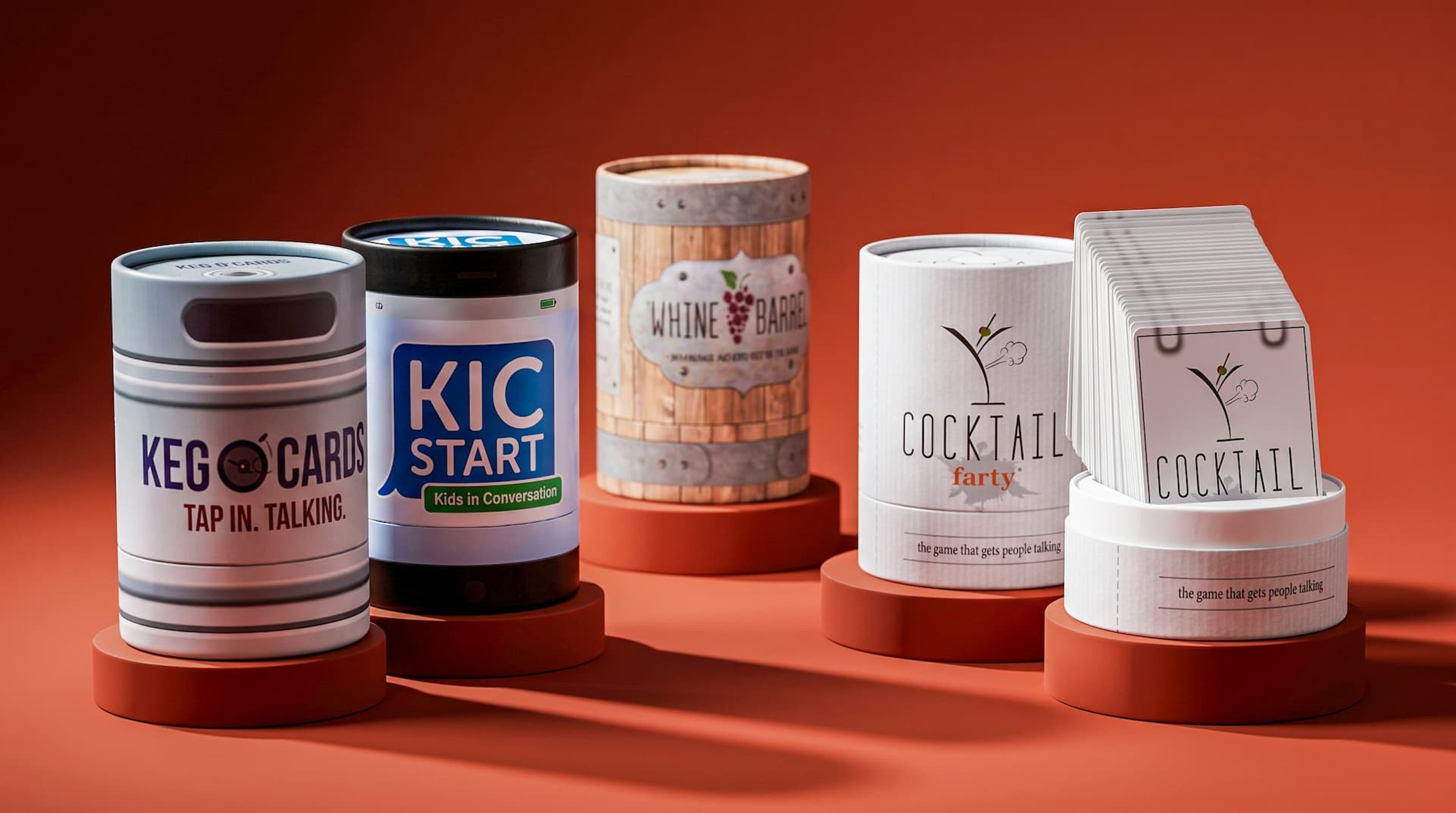 Five custom cylindrical game boxes on round risers against a red background, with the Cocktail Farty game open on the right.