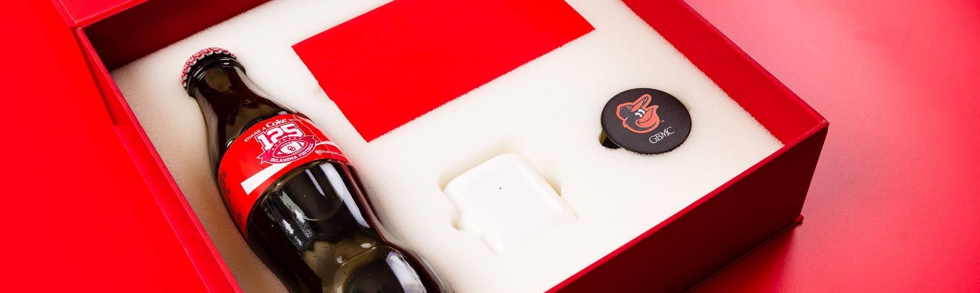 Close-up of an open Coca-Cola gift box holding a glass bottle, AirPods case, and PopSocket in foam.
