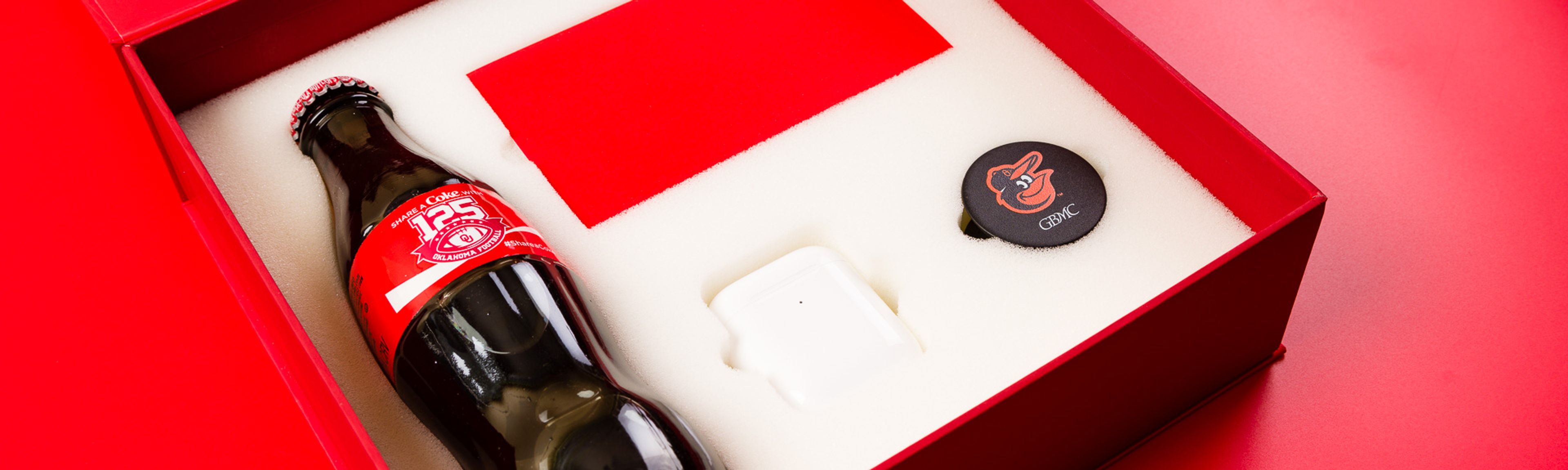Close-up of an open Coca-Cola gift box holding a glass bottle, AirPods case, and PopSocket in foam.