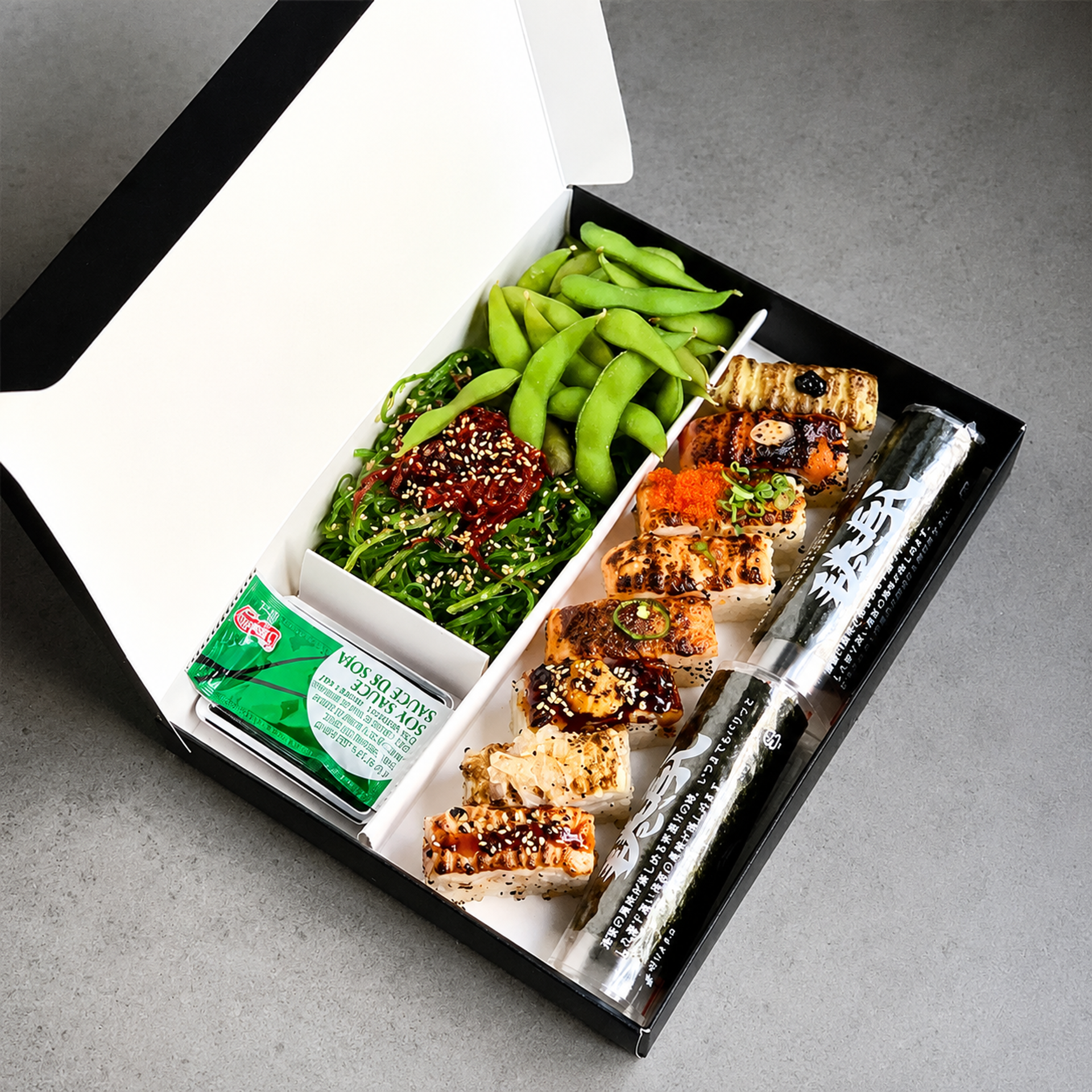 Overhead view of an open Torch Sushi bento box with aburi sushi, edamame, seaweed salad, soy sauce, and a handroll.