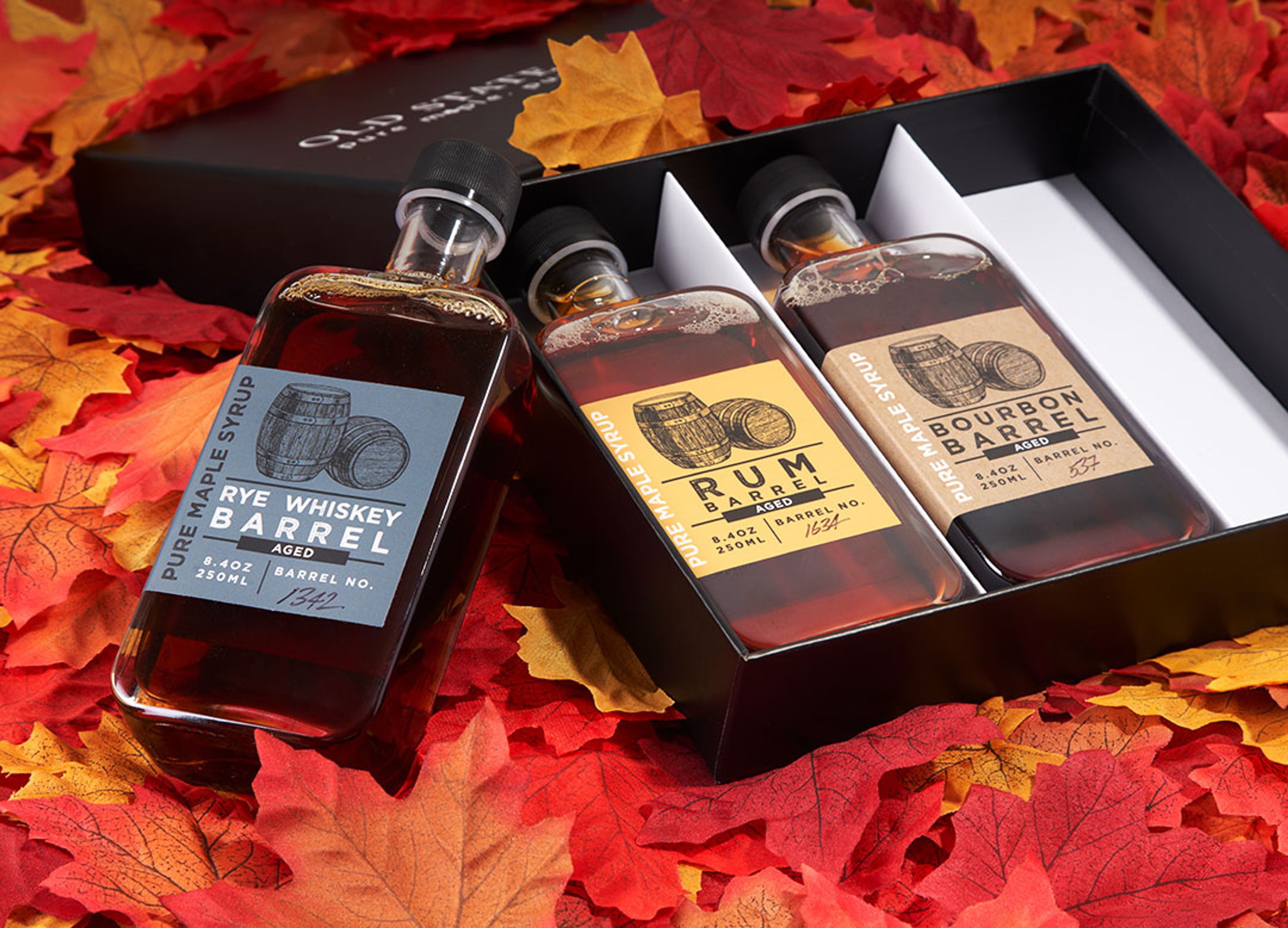Three bottles of Old State Farms barrel-aged syrup displayed in an open black rigid box surrounded by autumn leaves.