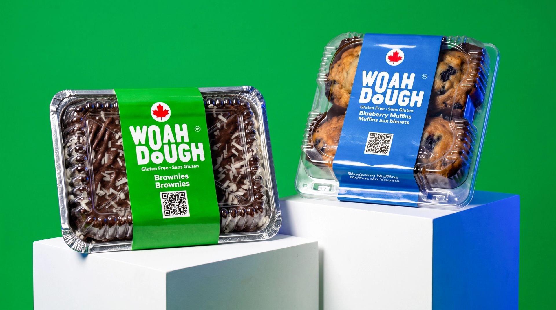 Woah Dough brownies and blueberry muffins in clear packaging with green and blue branded sleeves on a green backdrop.
