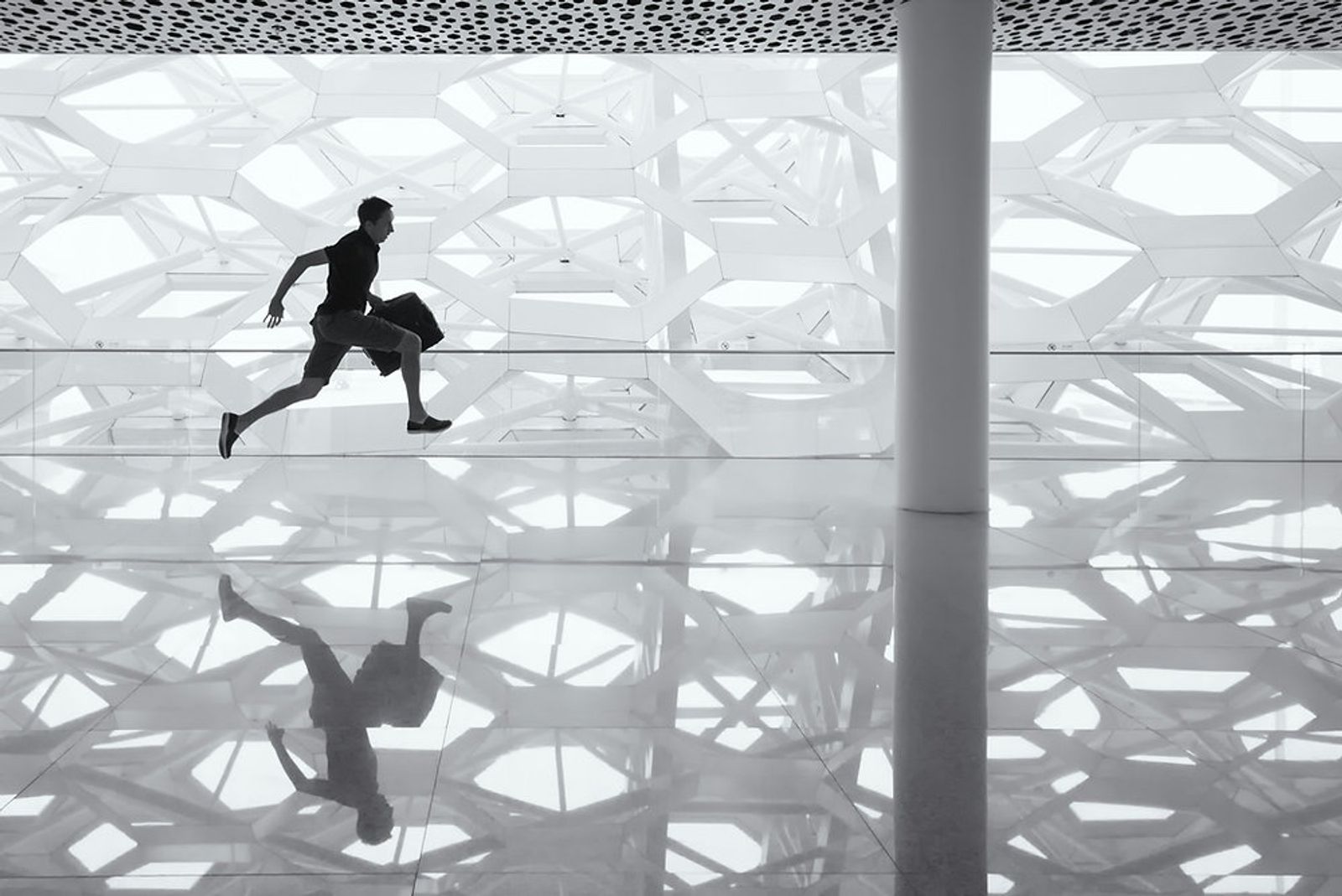 Man running with a bag past a white geometric lattice wall, mirrored in a polished floor