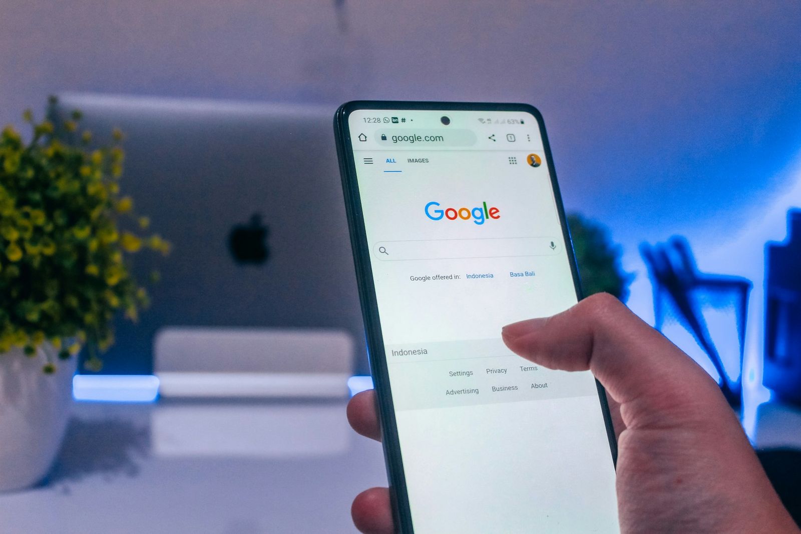 Hand holding a smartphone with Google's search homepage open, in front of an iMac