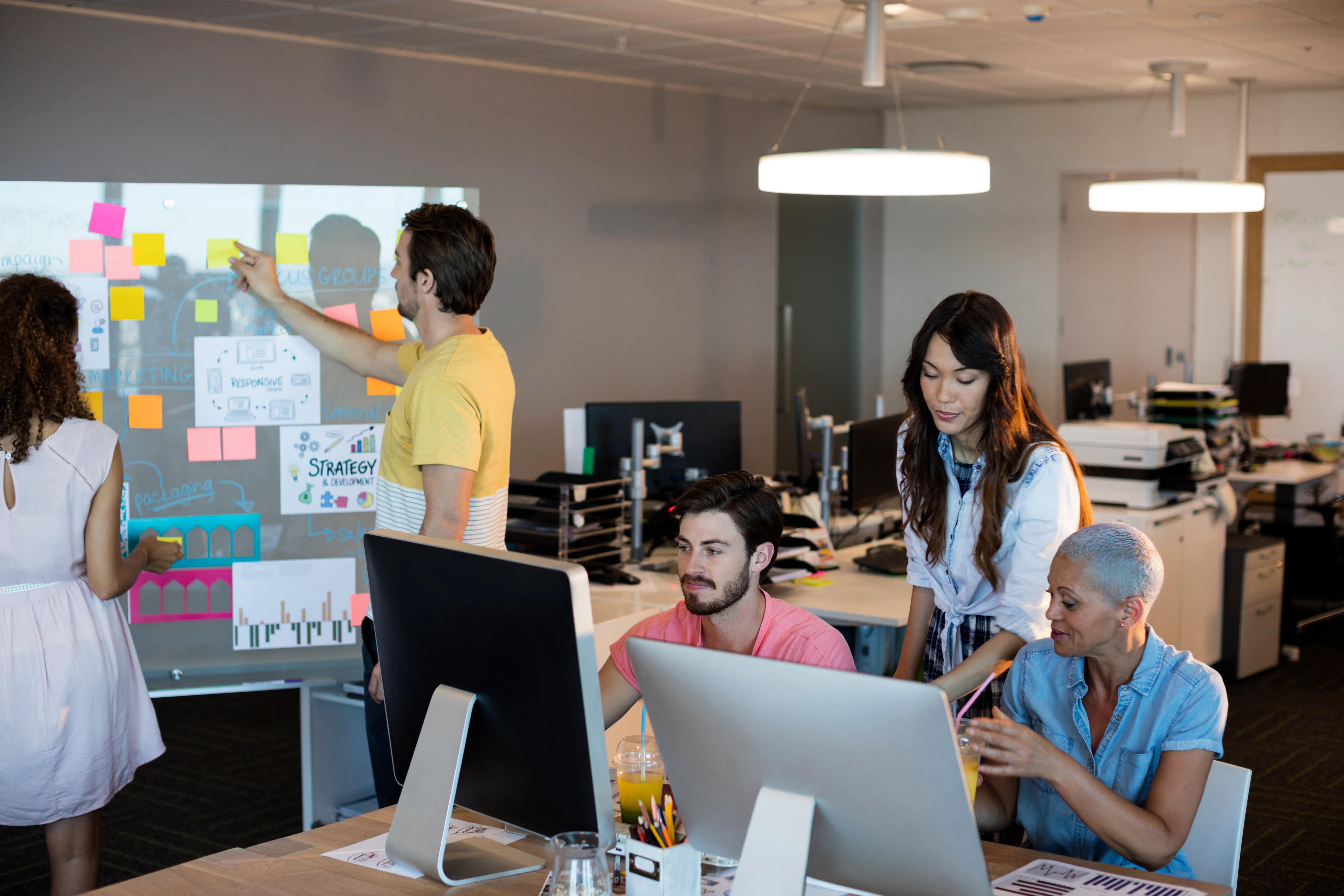 A diverse creative business team actively engaged in a strategy session with one member pointing at sticky notes on a whiteboard, in a dynamic office workspace.