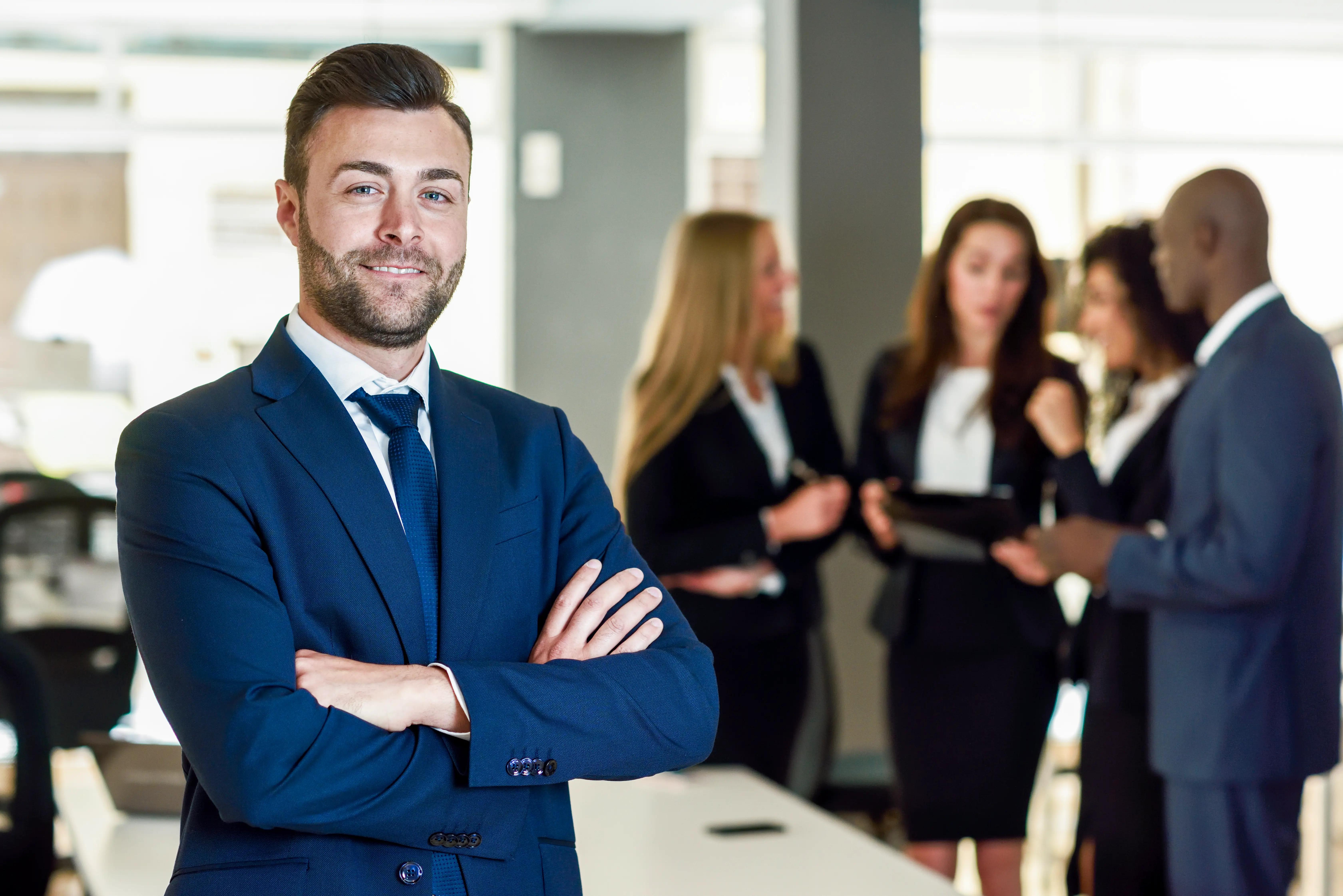 Confident businessman with crossed arms standing in the foreground, with a team of professionals discussing in the background of a modern office, representing leadership in accounting services.