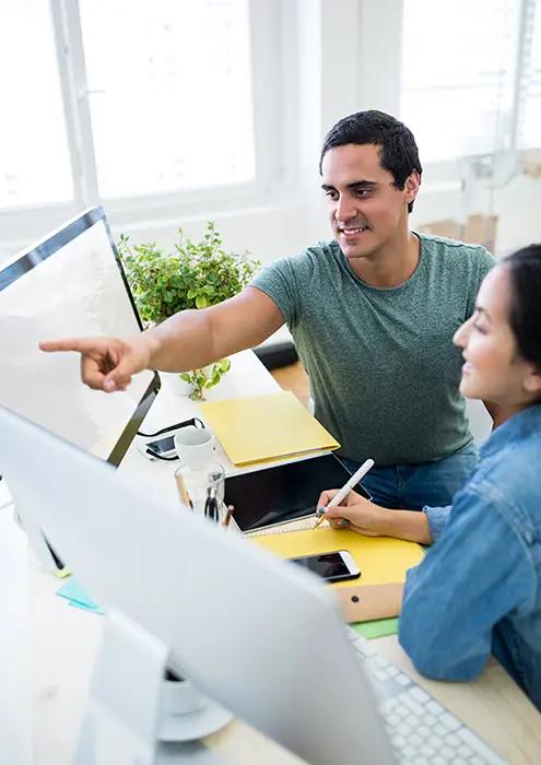 A male web developer in a green t-shirt discussing project details with a female colleague over a computer screen in a bright office, representing a collaborative environment in web development services.