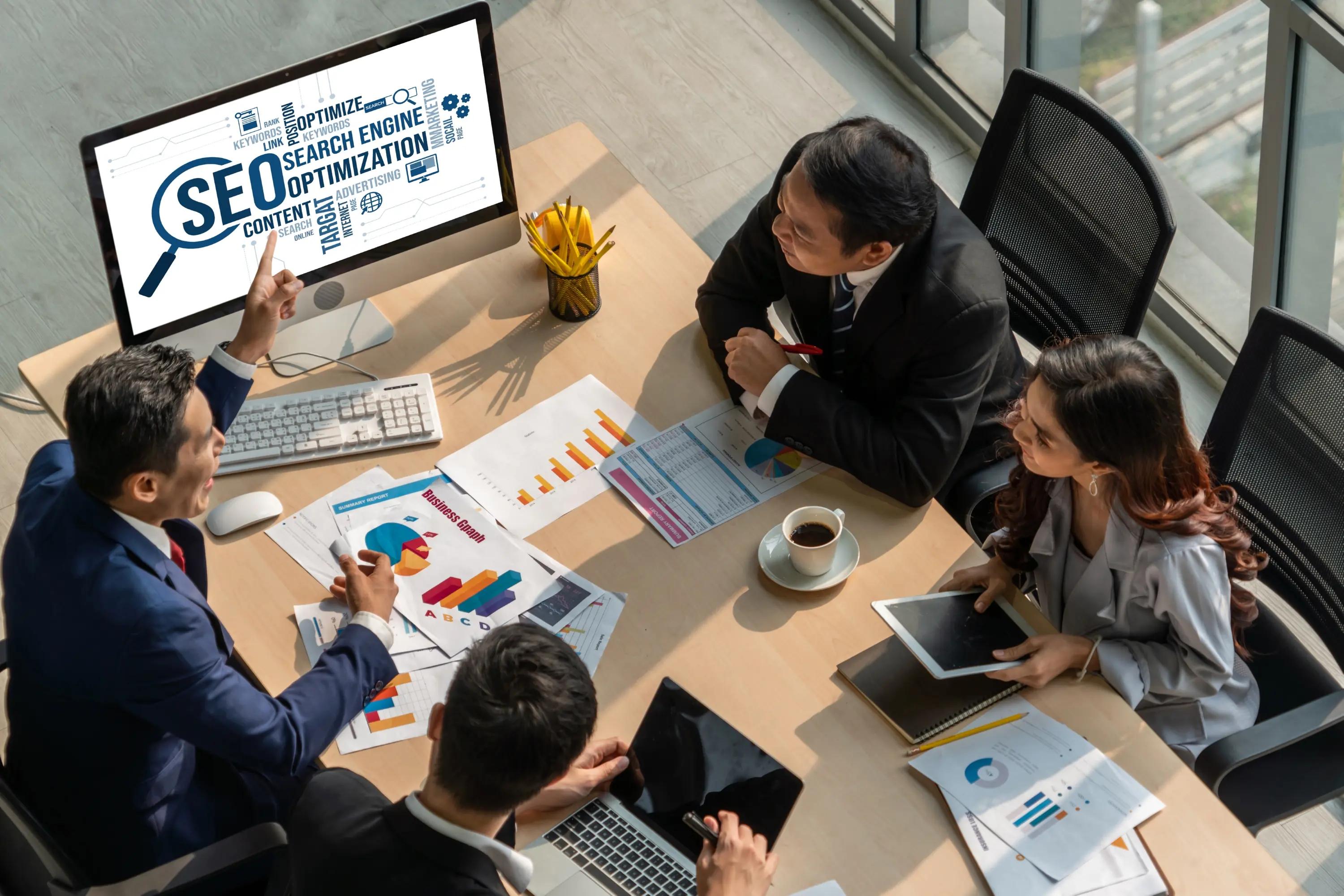 A business team engaged in a strategic meeting with a focus on SEO, as evidenced by a large monitor displaying 'Search Engine Optimization' concepts, in a contemporary office setting