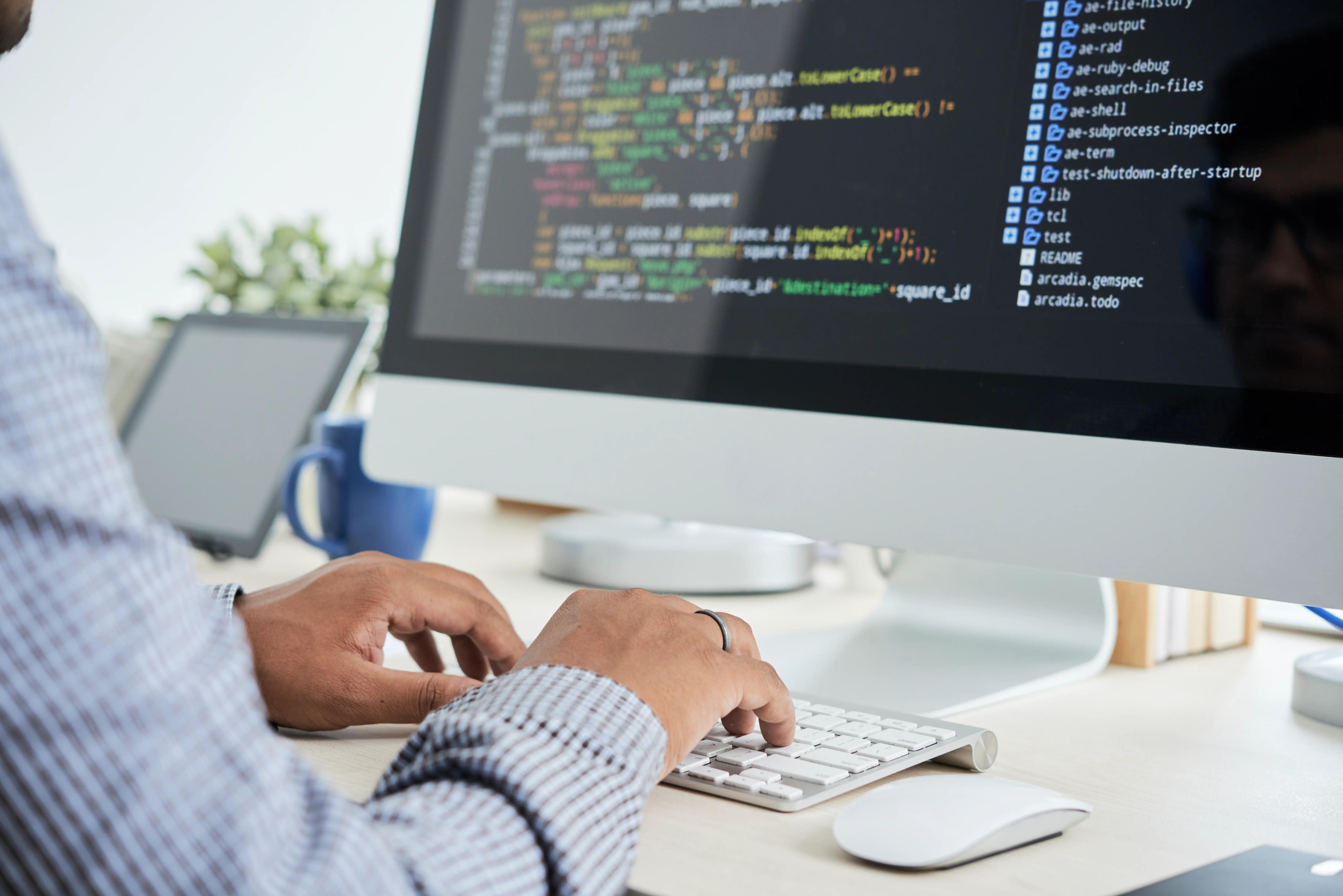 Professional web developer providing web development services, typing on a keyboard with coding software on a computer screen displaying code in a bright office setting