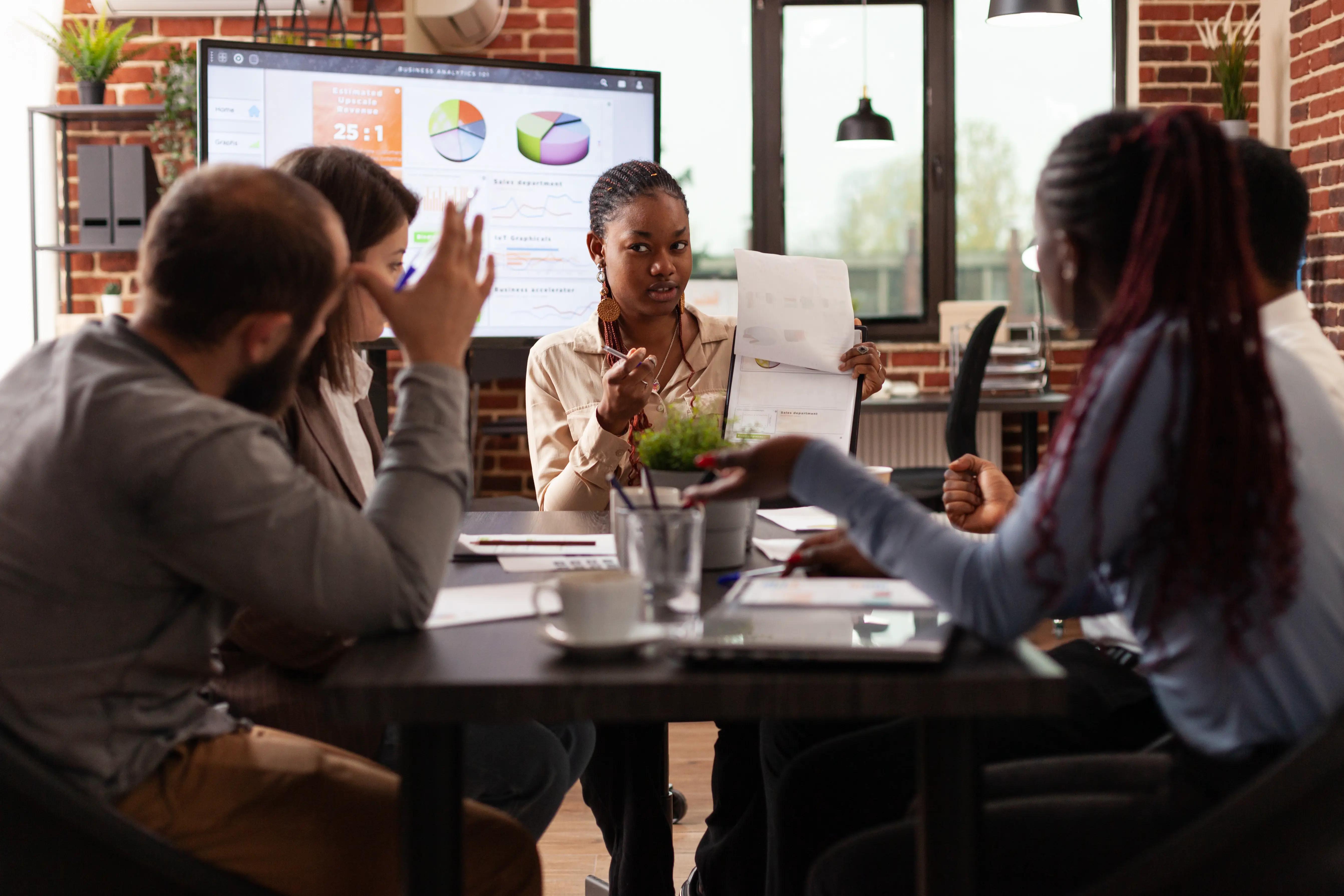 A diverse business team is engaged in a lively discussion over documents and a presentation with analytics and pie charts on a screen, in a modern brick-walled office.