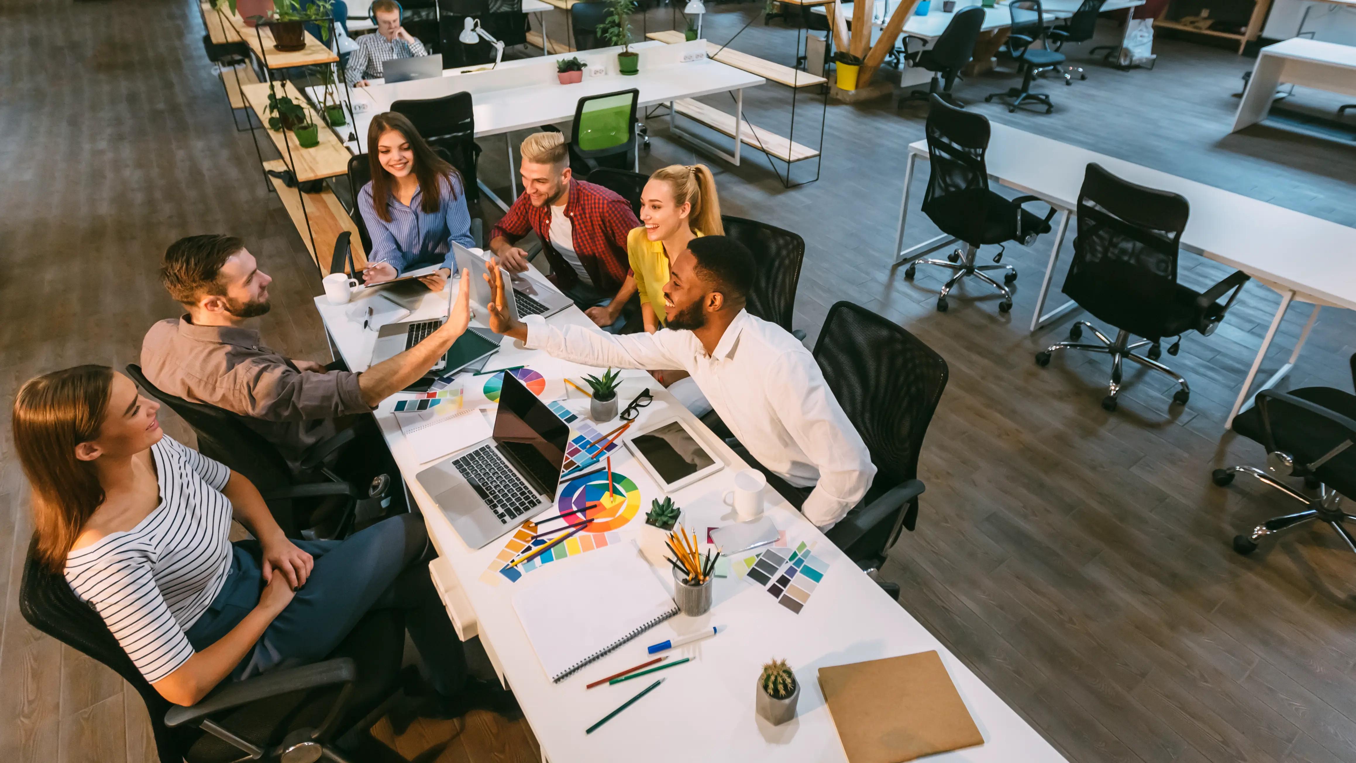 A group of creative professionals are gathered around a table in a vibrant office setting, engaged in a celebratory moment with a high-five. The workspace is well-equipped with laptops, color swatches, and design tools, suggesting a collaborative and dynamic work environment. The diverse team seems to be in high spirits, possibly celebrating a successfully completed project or a creative breakthrough