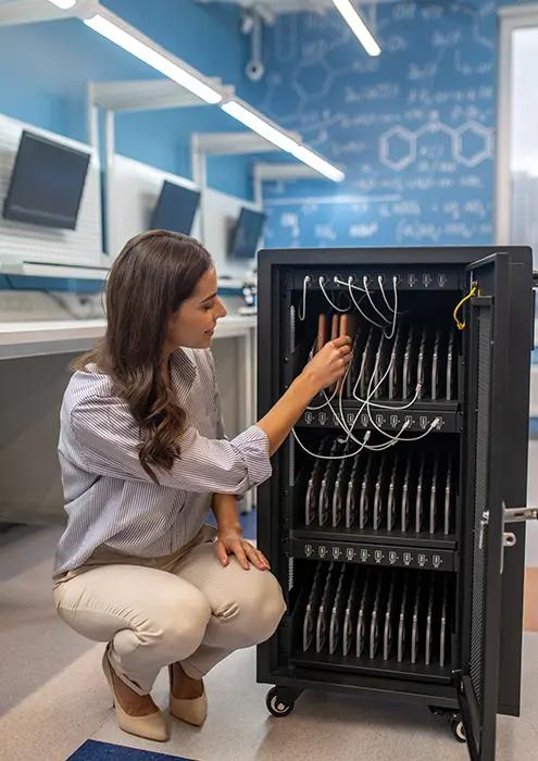 A woman in business casual attire crouching and working on a server rack cabinet in a server room with computer workstations and digital blueprints in the background.