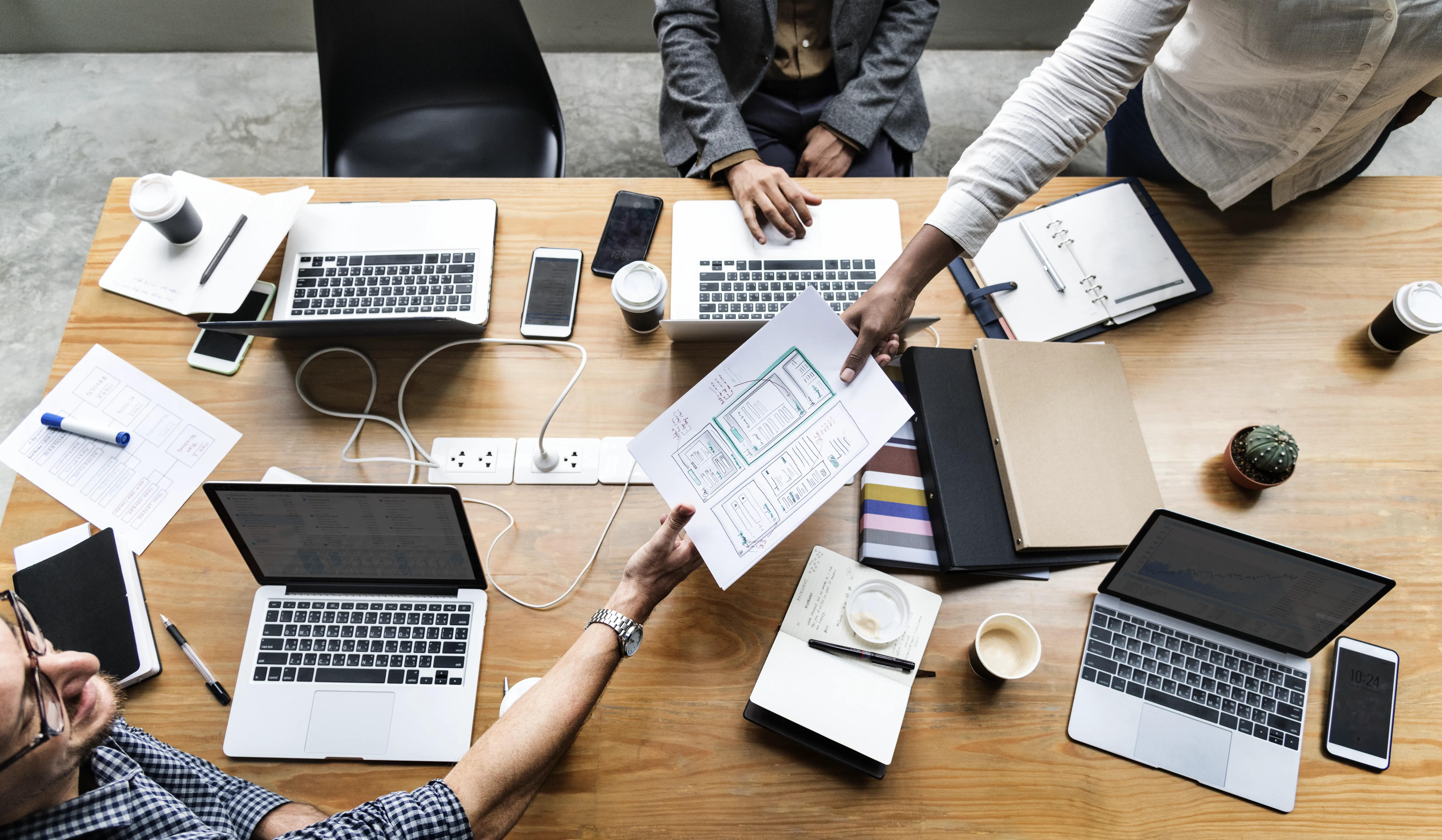 Top view of a collaborative team meeting with members engaged in app development services, surrounded by laptops, mobile devices, and UI/UX design layouts on a wooden table in a modern workspace