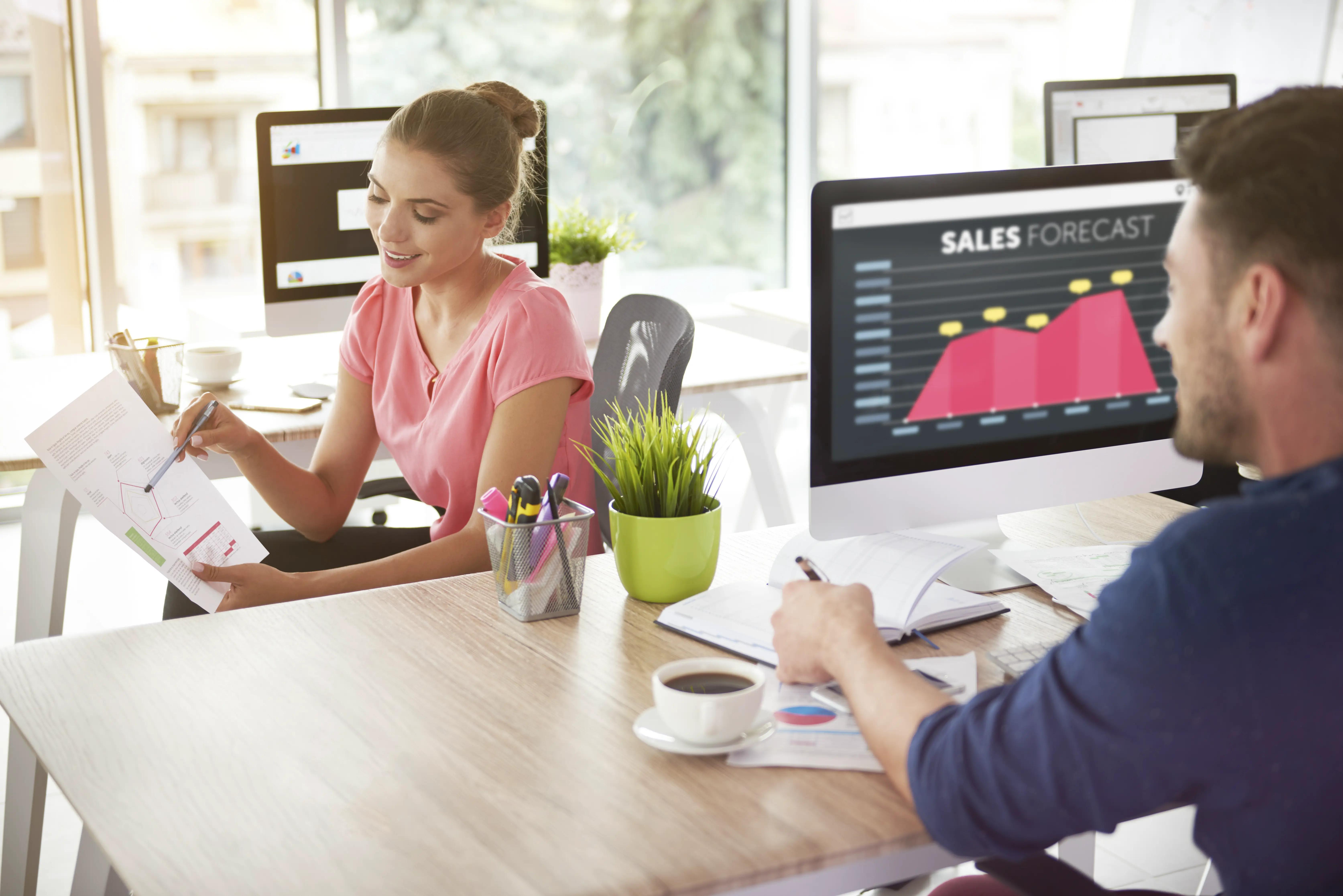 Two colleagues, a woman and a man, are analyzing documents in a bright office environment, with a 'Sales Forecast' graph visible on a computer monitor in the background