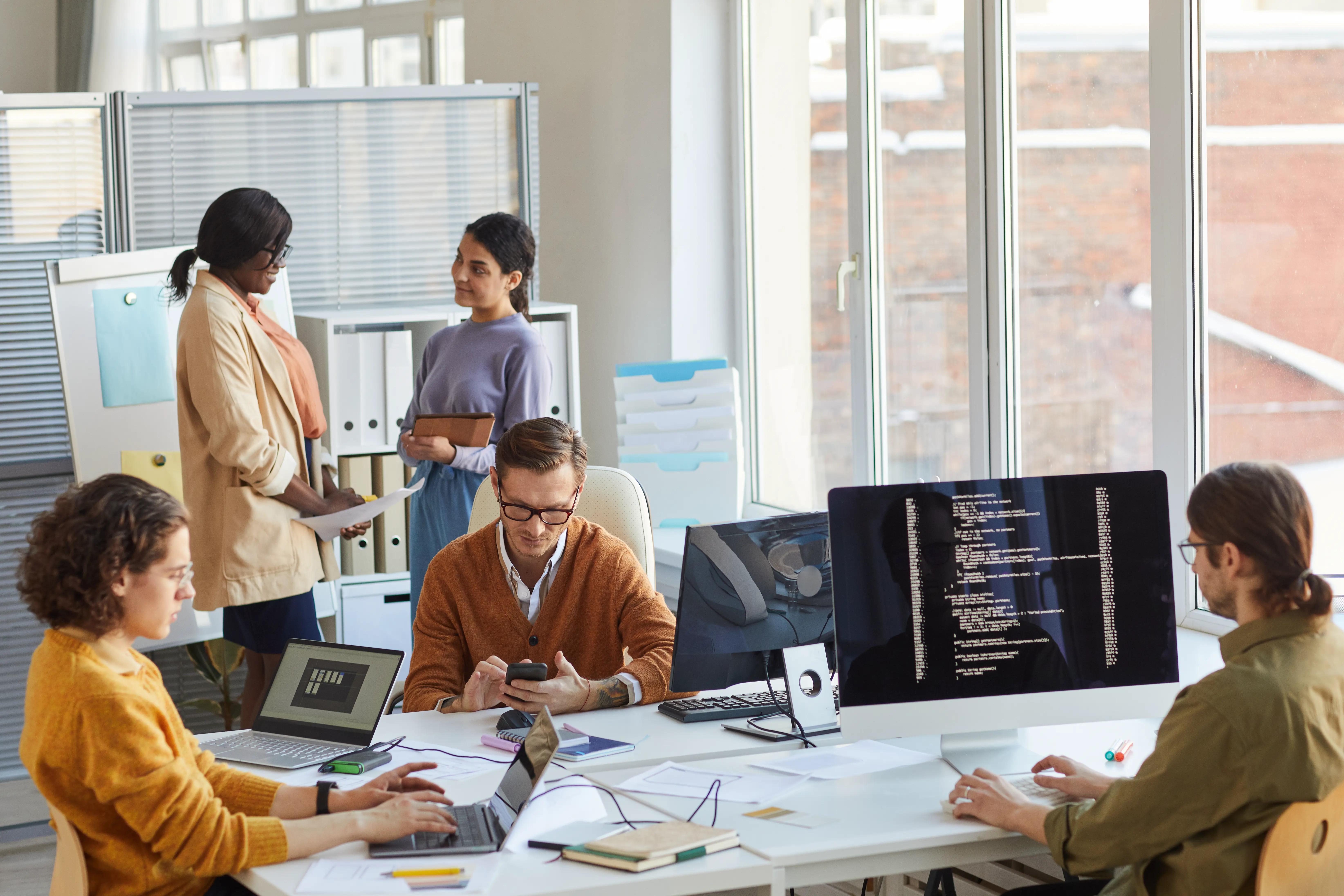 Diverse team of IT developers collaborating on a project in a software production studio, with computers displaying programming code, indicative of professional web development services.