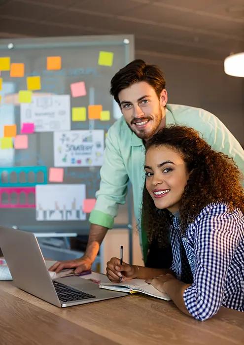 A man and woman team collaborating over a laptop with a colorful brainstorming whiteboard in the background, indicative of a vibrant, idea-generating work environment.