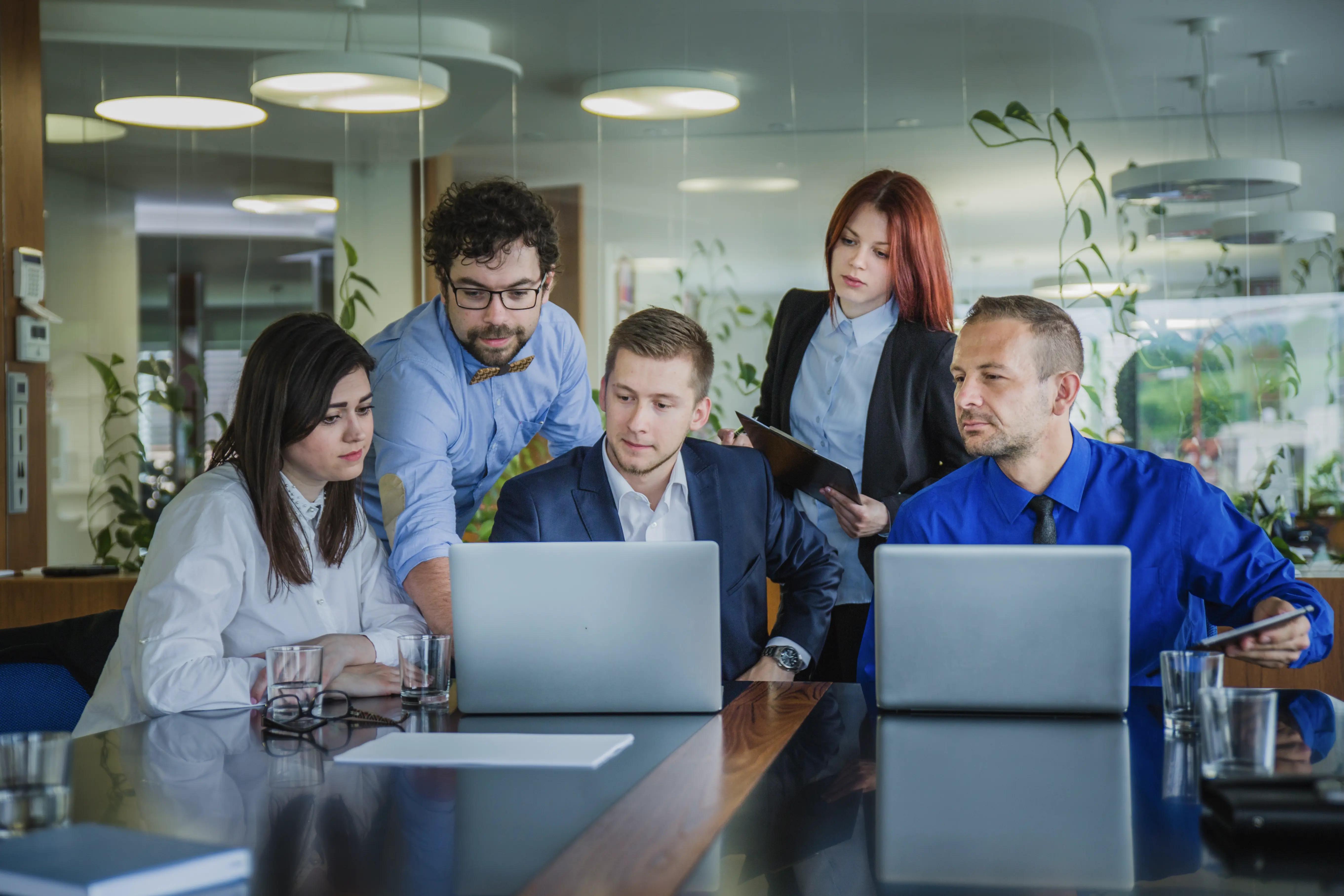 Focused team of professionals in a corporate meeting, working closely on laptops, likely discussing strategies for web development services in a modern office environment