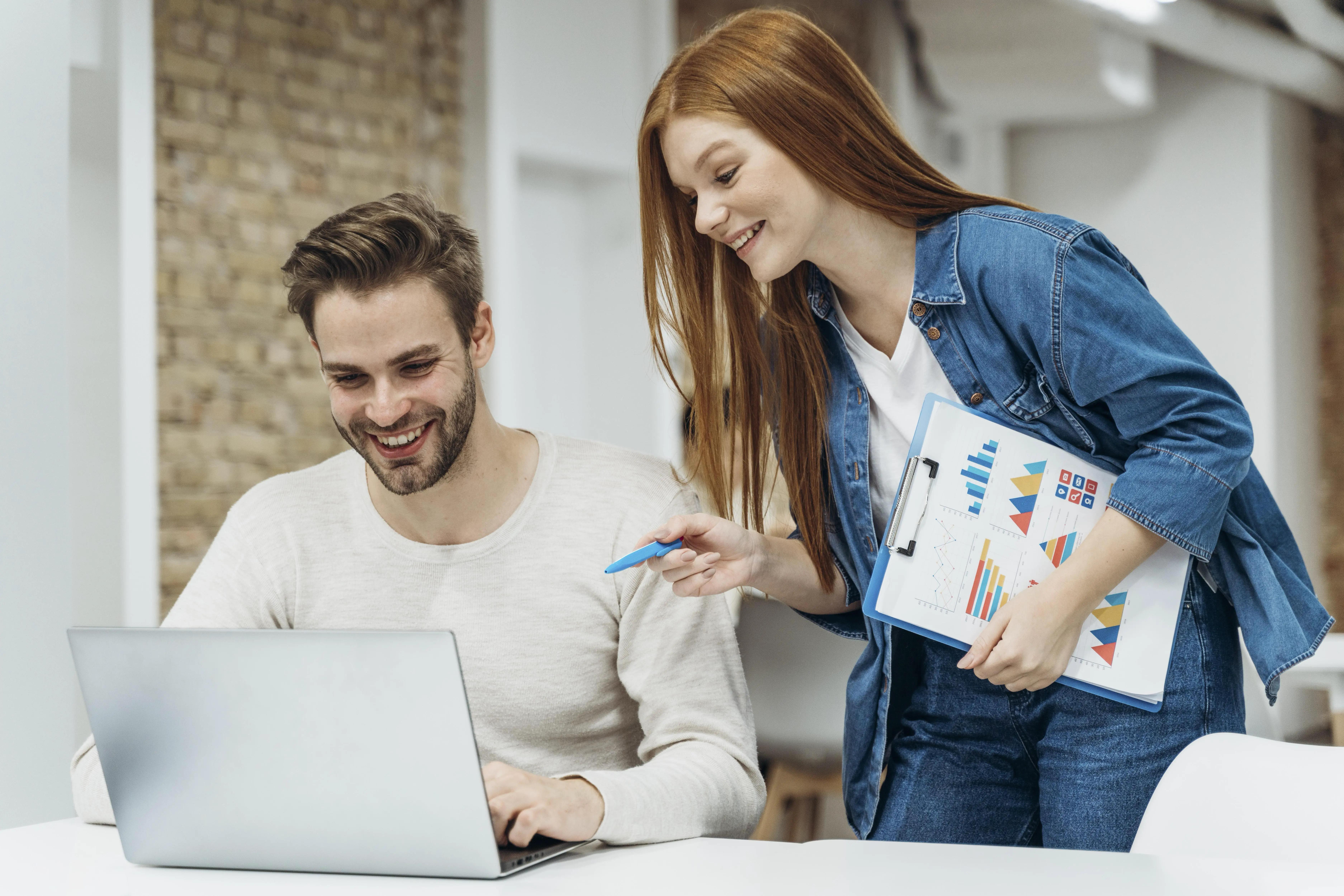 Two colleagues, a man and a woman, discussing a business project with the woman holding a clipboard with charts, indicative of a PPC marketing strategy session in a modern office.