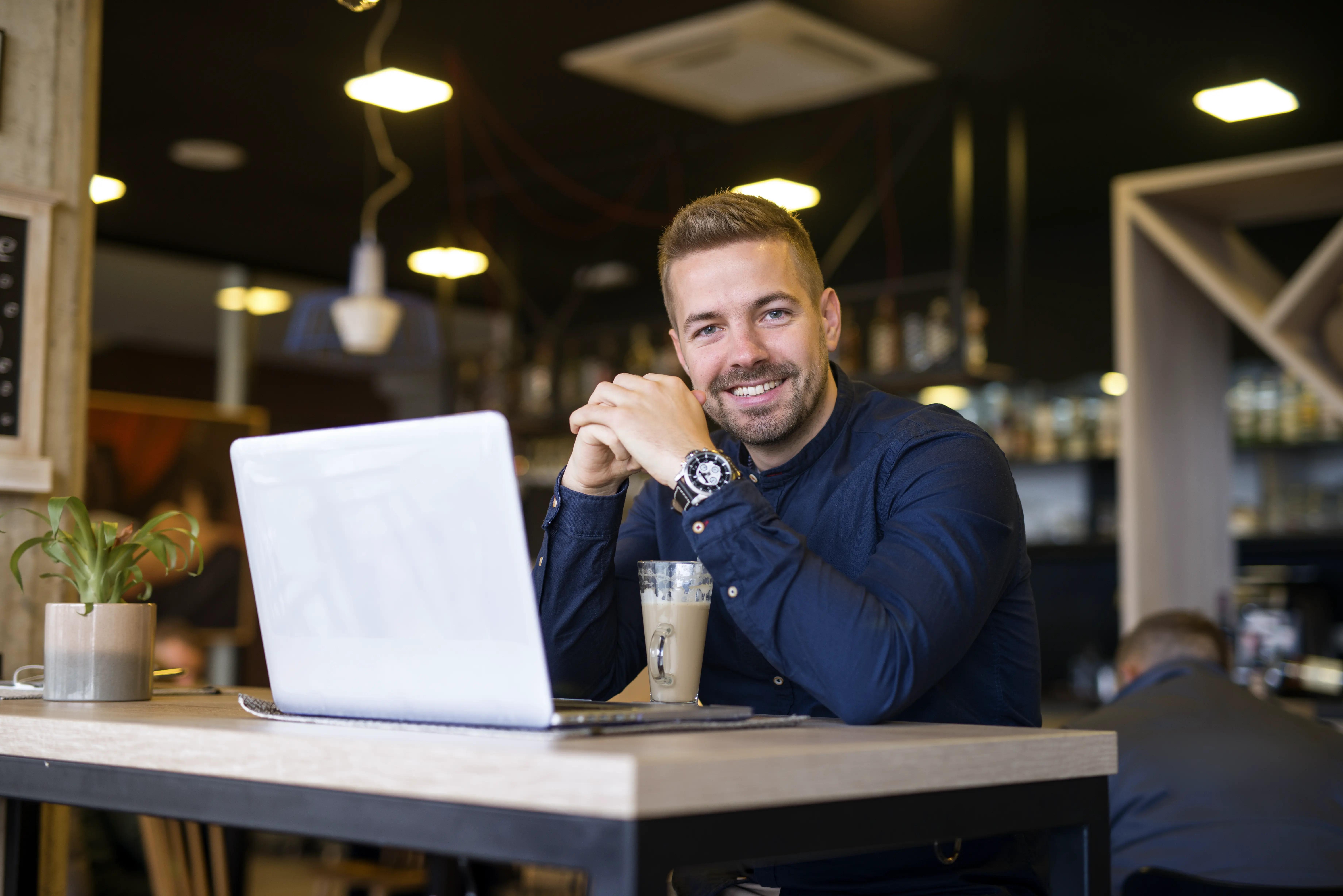 Portrait of a smiling man sitting at a café with his laptop, a casual yet productive setting, with a warm ambiance and a coffee on the table.