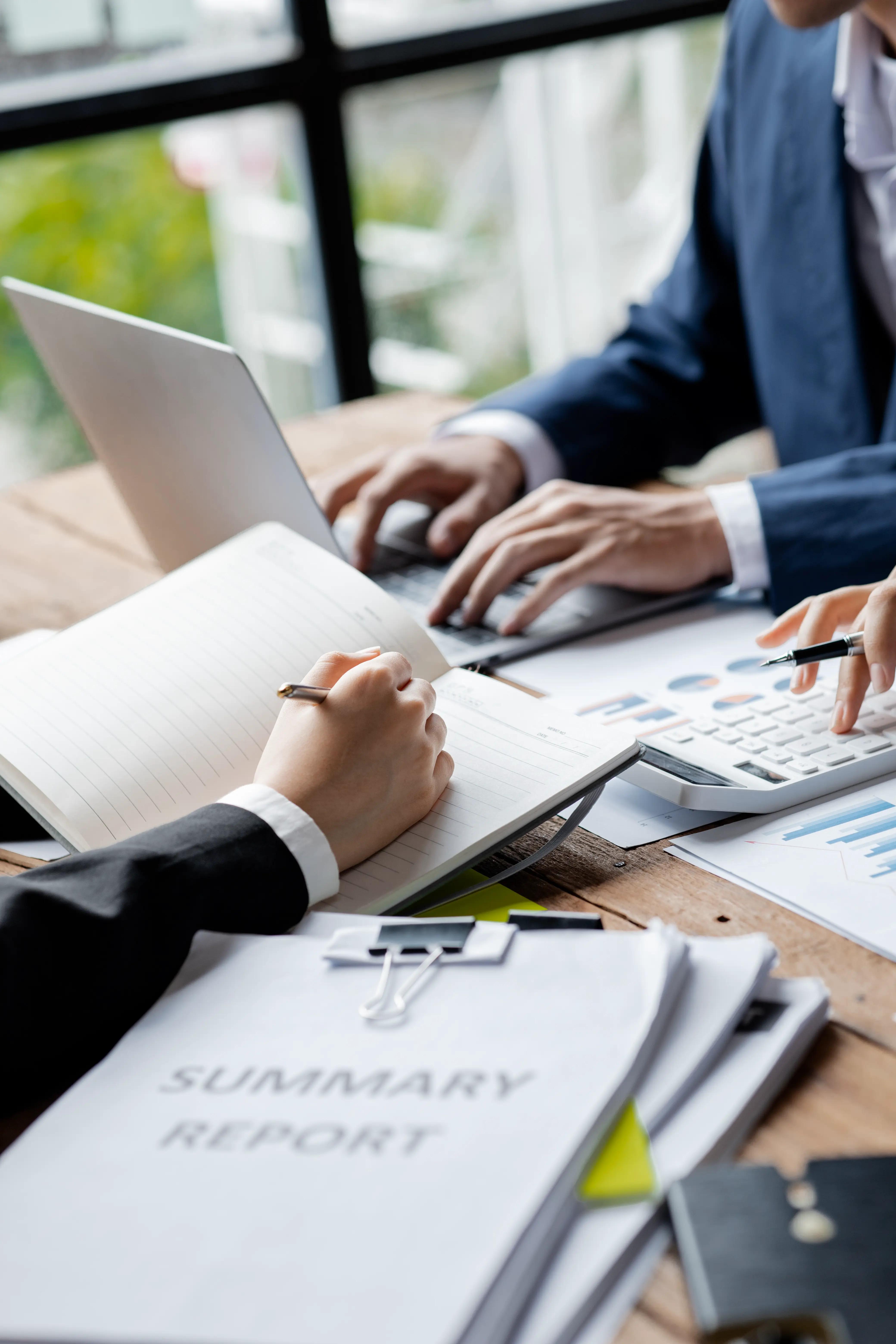 Close-up of business professionals working on financial reports, with a focus on hands writing in a notebook and using a calculator, suggesting meticulous accounting services