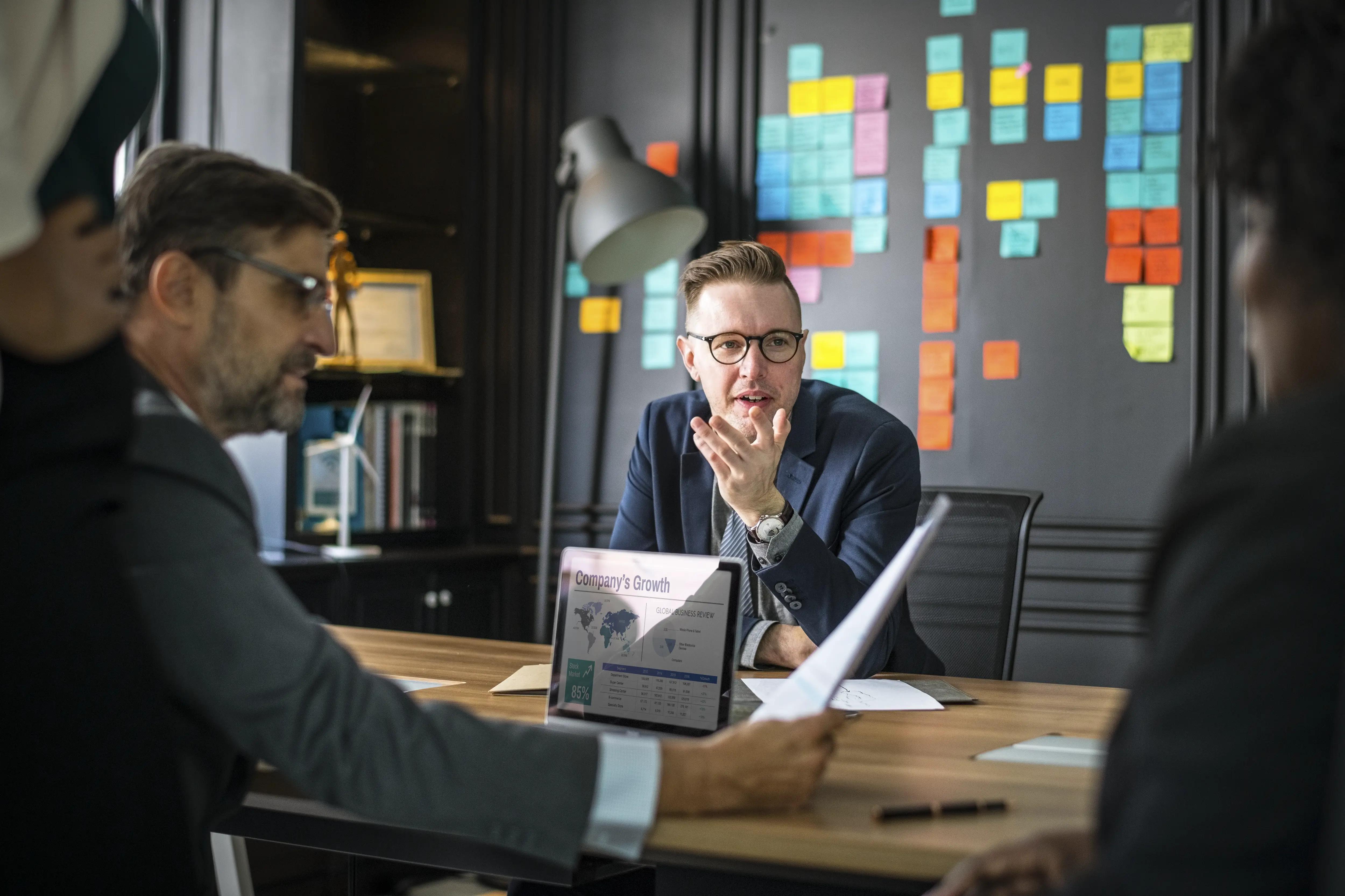 Engaged business team in a strategic meeting, with focus on a male leader explaining growth concepts, accompanied by colorful sticky notes on a board in a well-lit modern office setting.