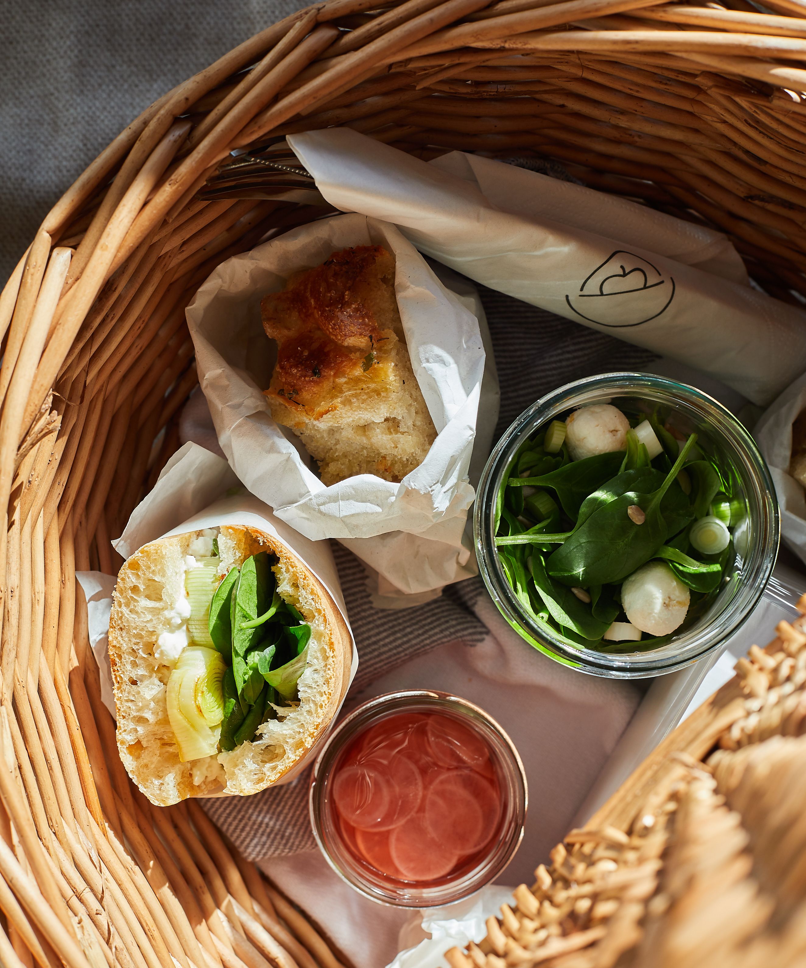 Picknickkorb gefüllt mit Ciabatta-Sandwich, Focaccia, Salat im Glas, eingelegten Radieschenscheiben im Glas