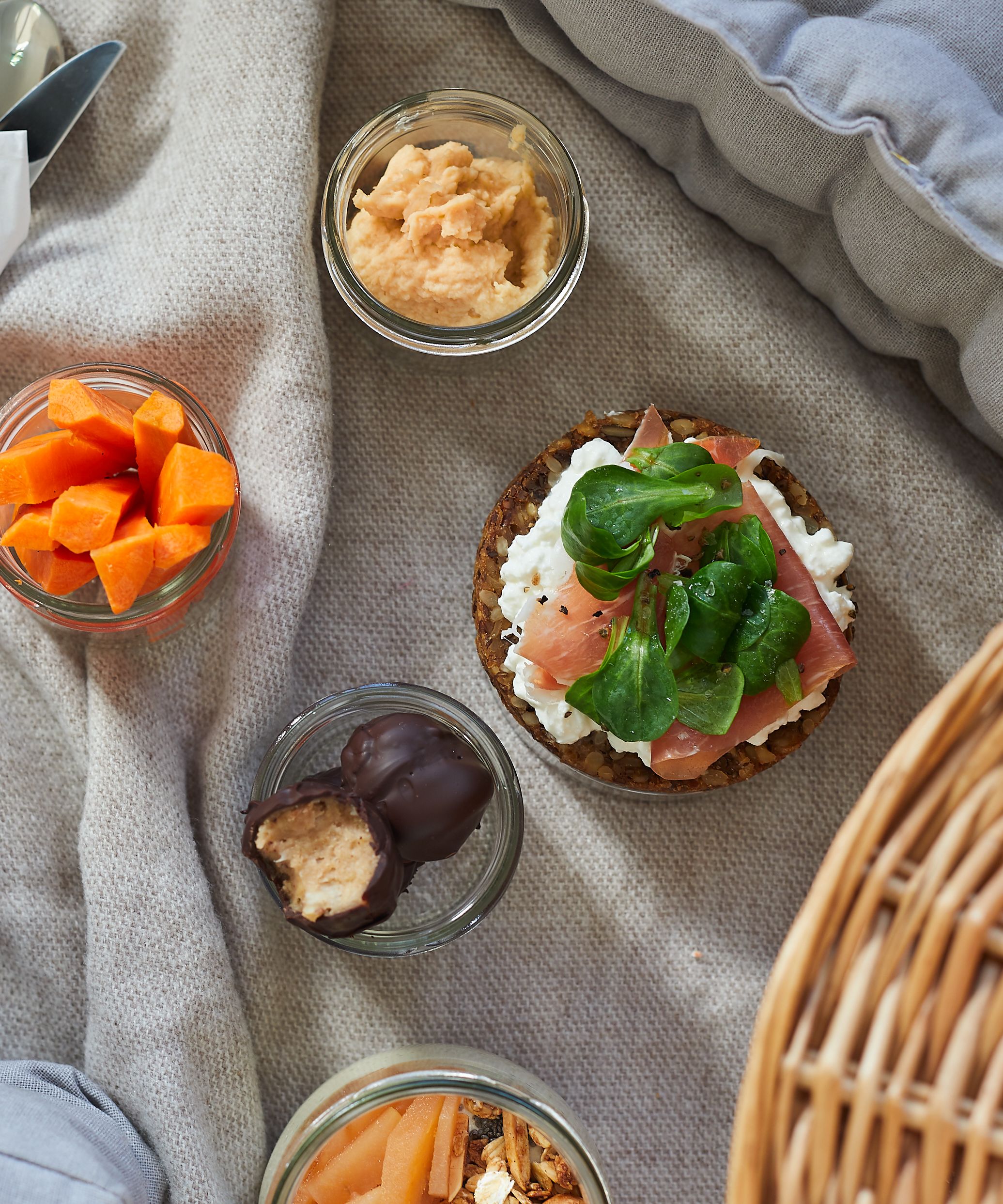 Picknickdecke mit geschnittenen Möhren, Cashew-Schokoladen-Kugeln, Hummus und Körnerbrot belegt mit körnigem Frischkäse, Schinken und Feldsalat
