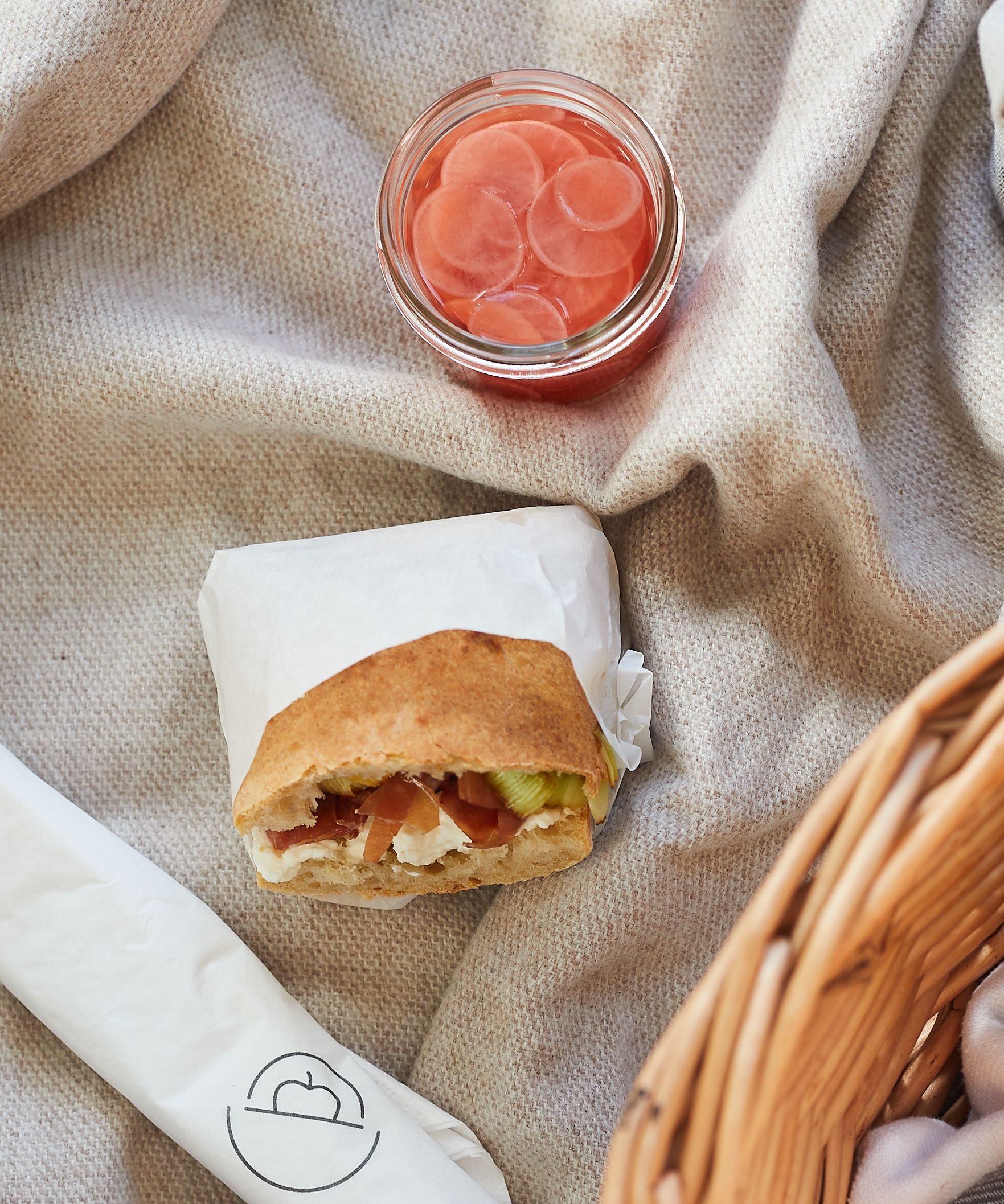 Ciabatta-Sandwich in Papier gewickelt, süß-saure Radieschen im Glas auf cremefarbener Decke