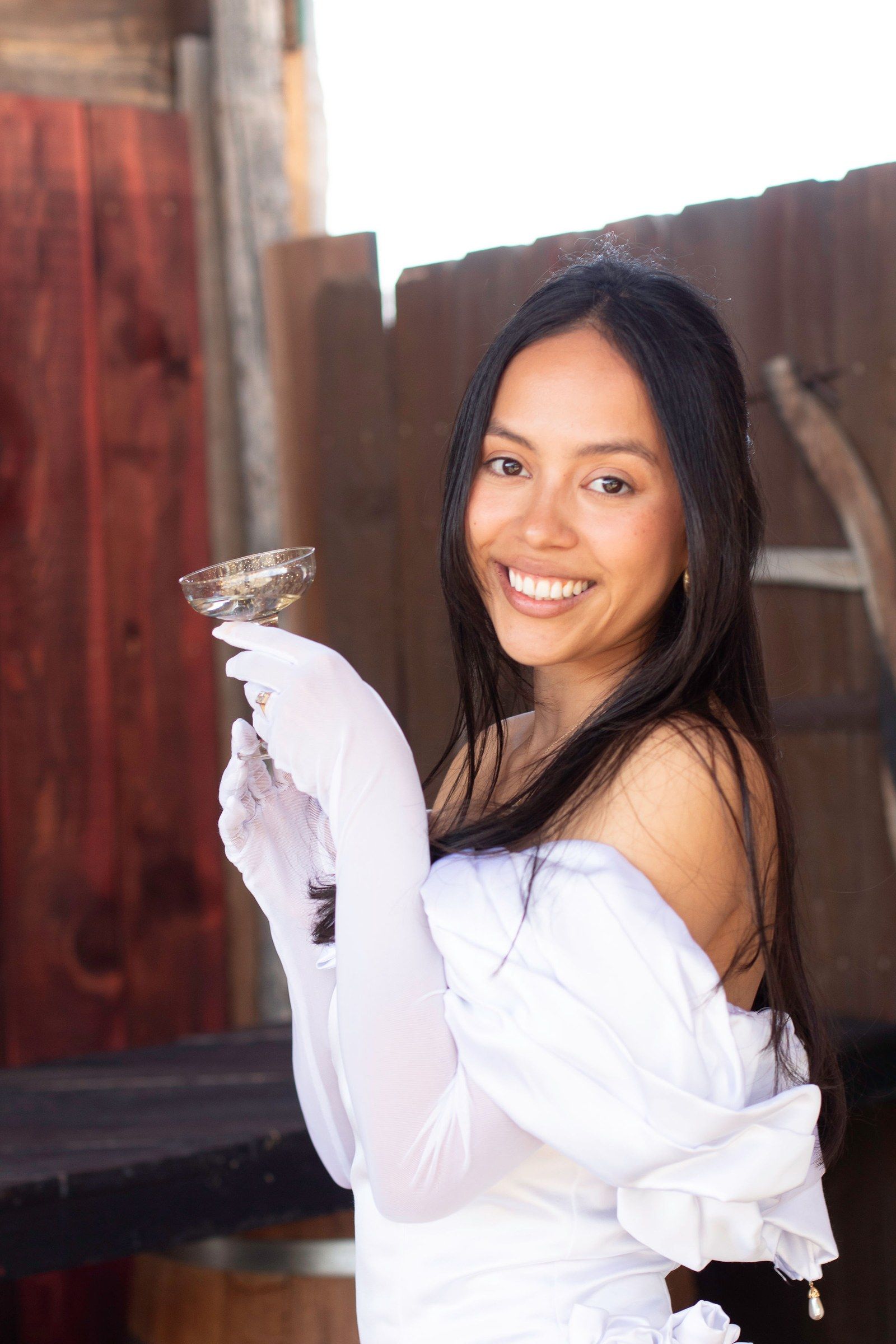 A bride in long white gloves raises a coupe of champagne against weathered red barn boards