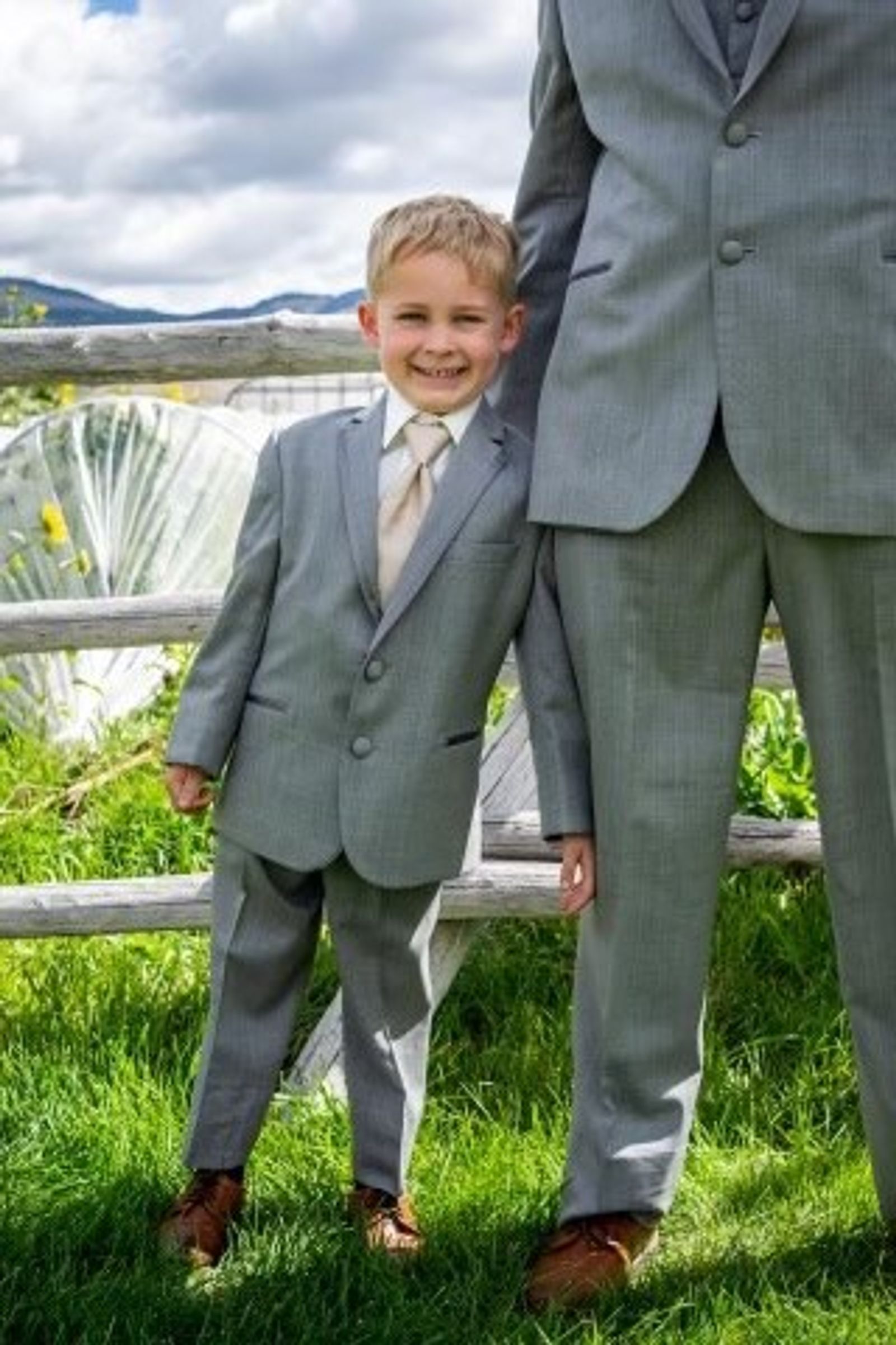 A young ring bearer in a gray suit and tie grins beside the jackleg fence