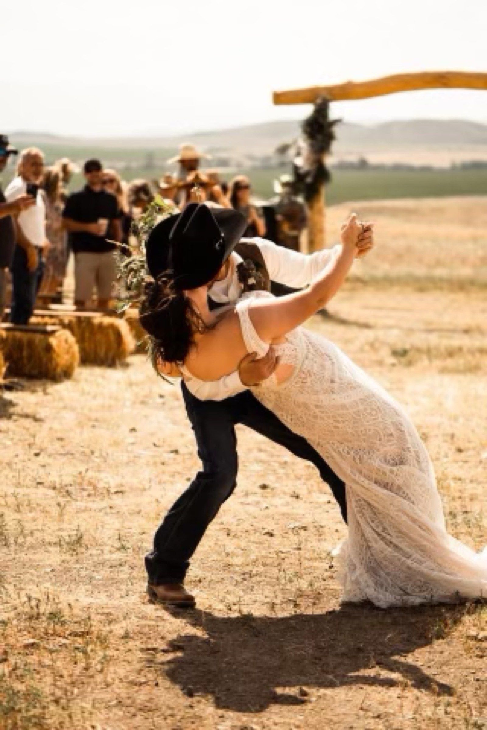 A groom in a black cowboy hat dips his bride as guests cheer beside the hay bales