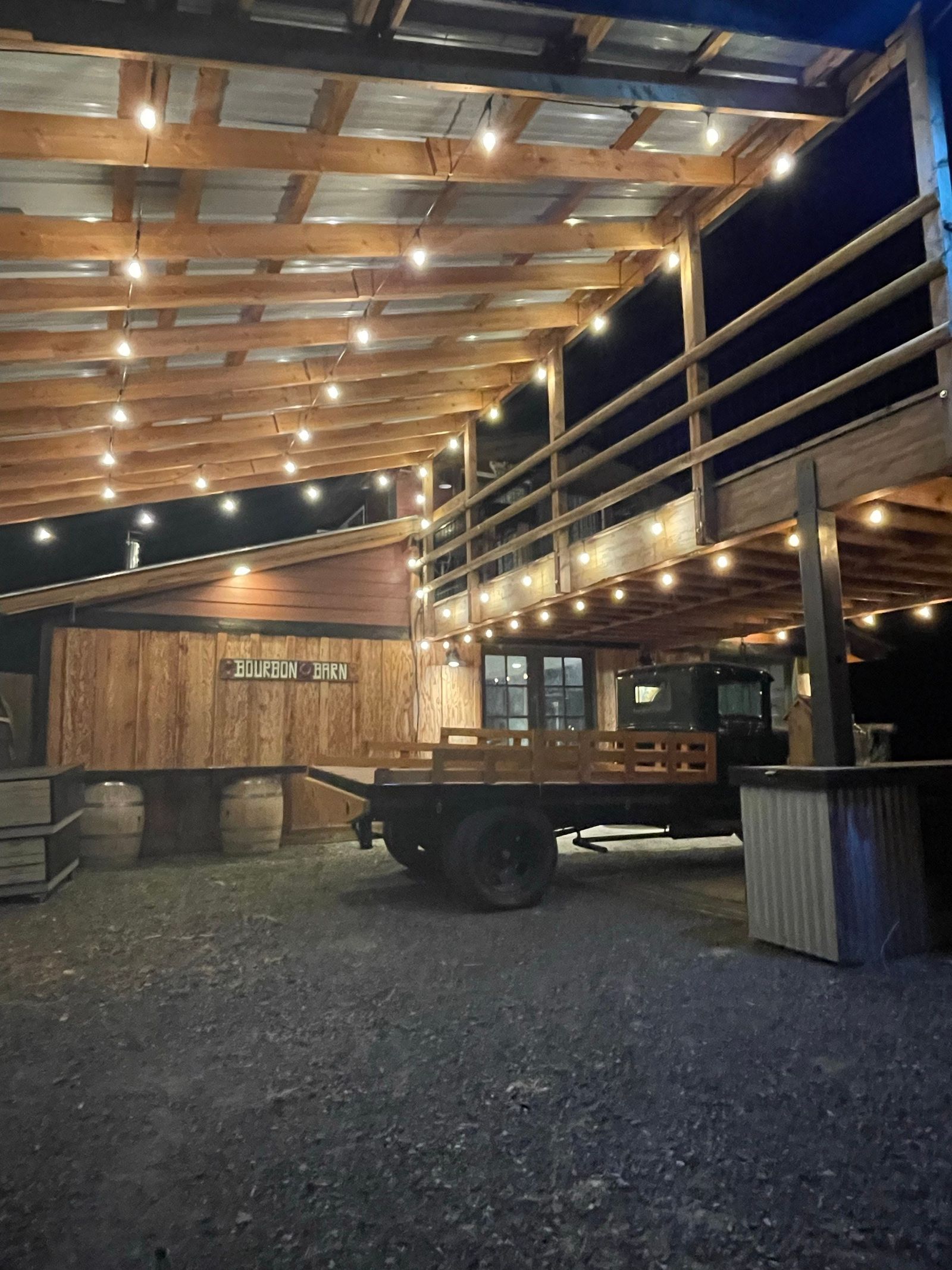 String lights glowing under the Bourbon Barn deck at night, the barn sign and the old flatbed beside the wall