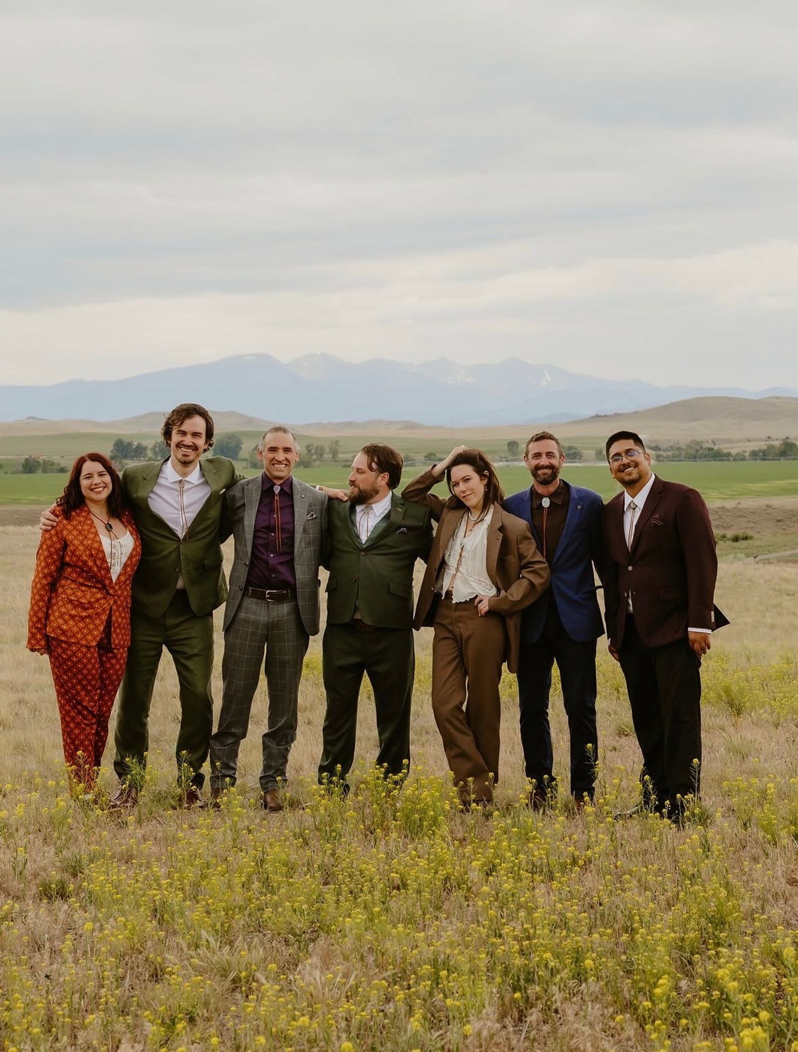 A wedding party in suits of every color standing arm in arm in the meadow, the mountains behind
