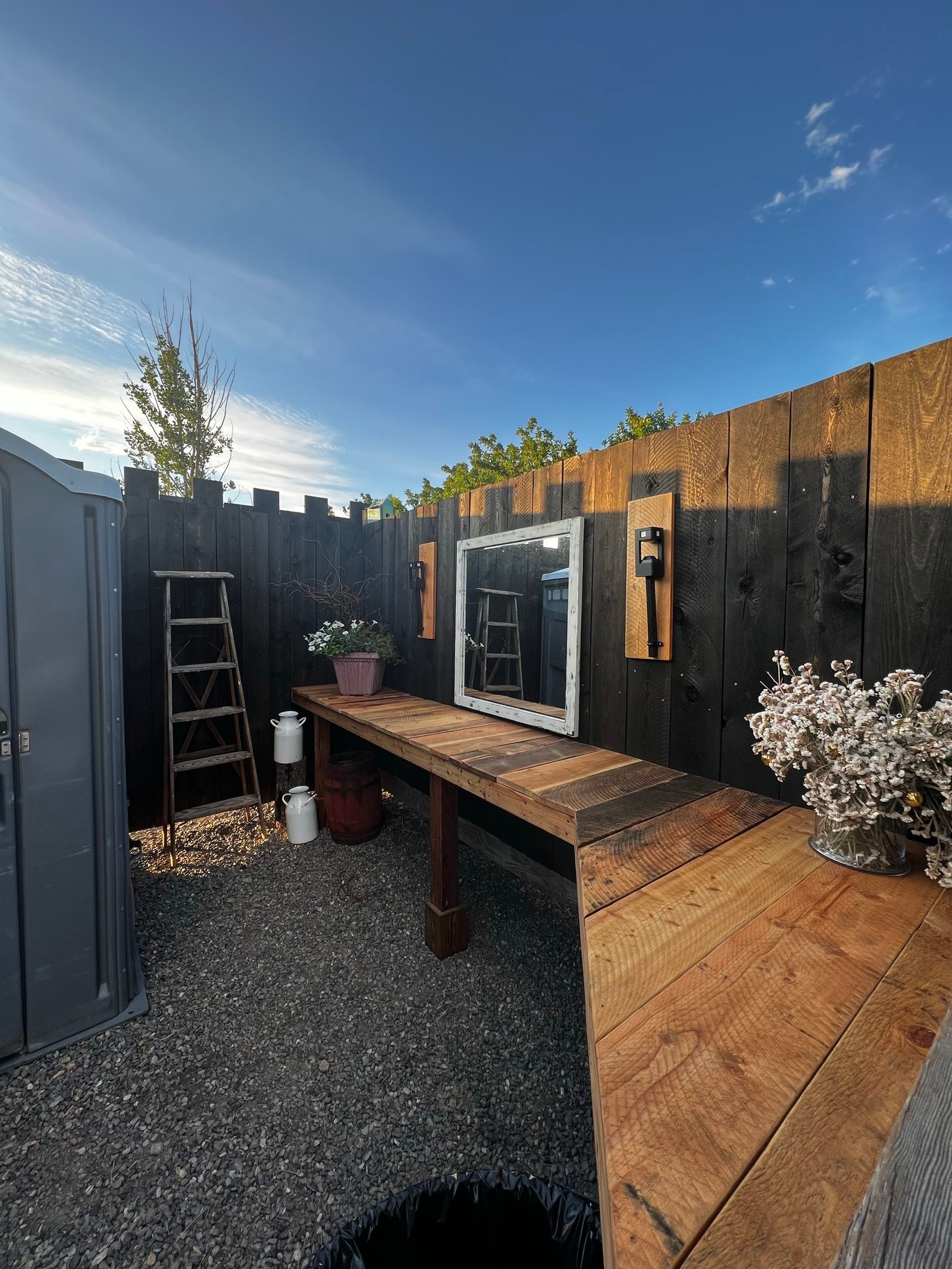 An outdoor wash station - a long wooden counter, mirror, and flowers against a black board fence