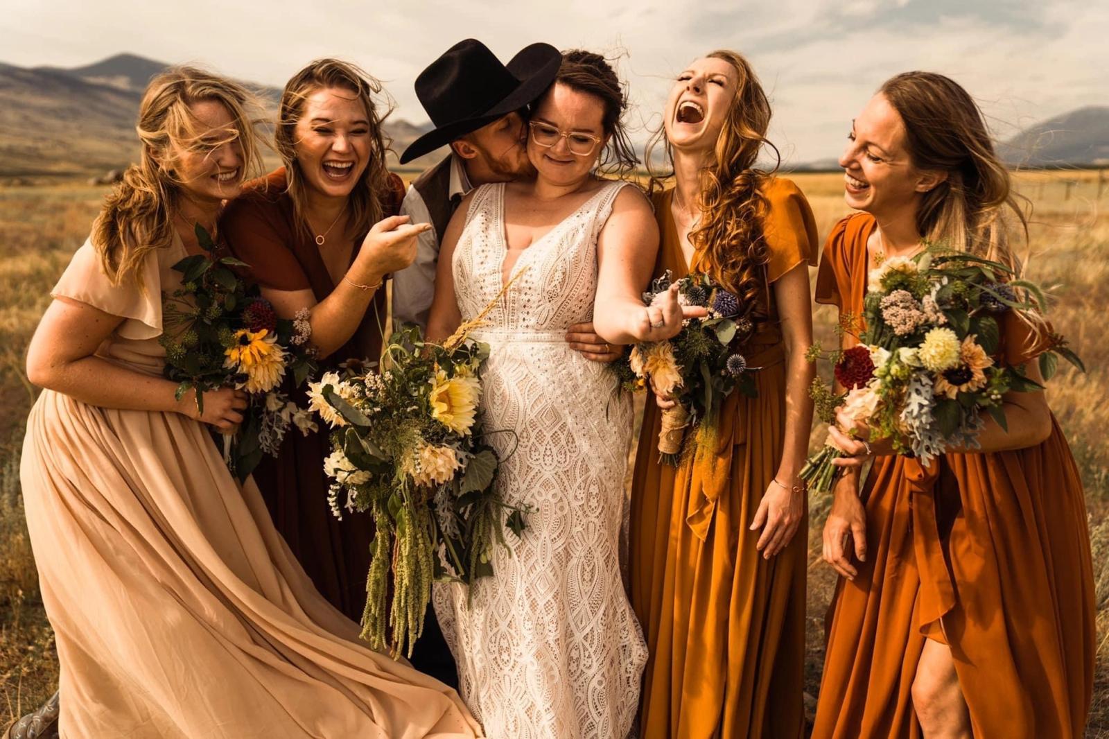A groom in a black cowboy hat kisses his bride's cheek as her bridesmaids in rust and blush dresses laugh in the prairie wind