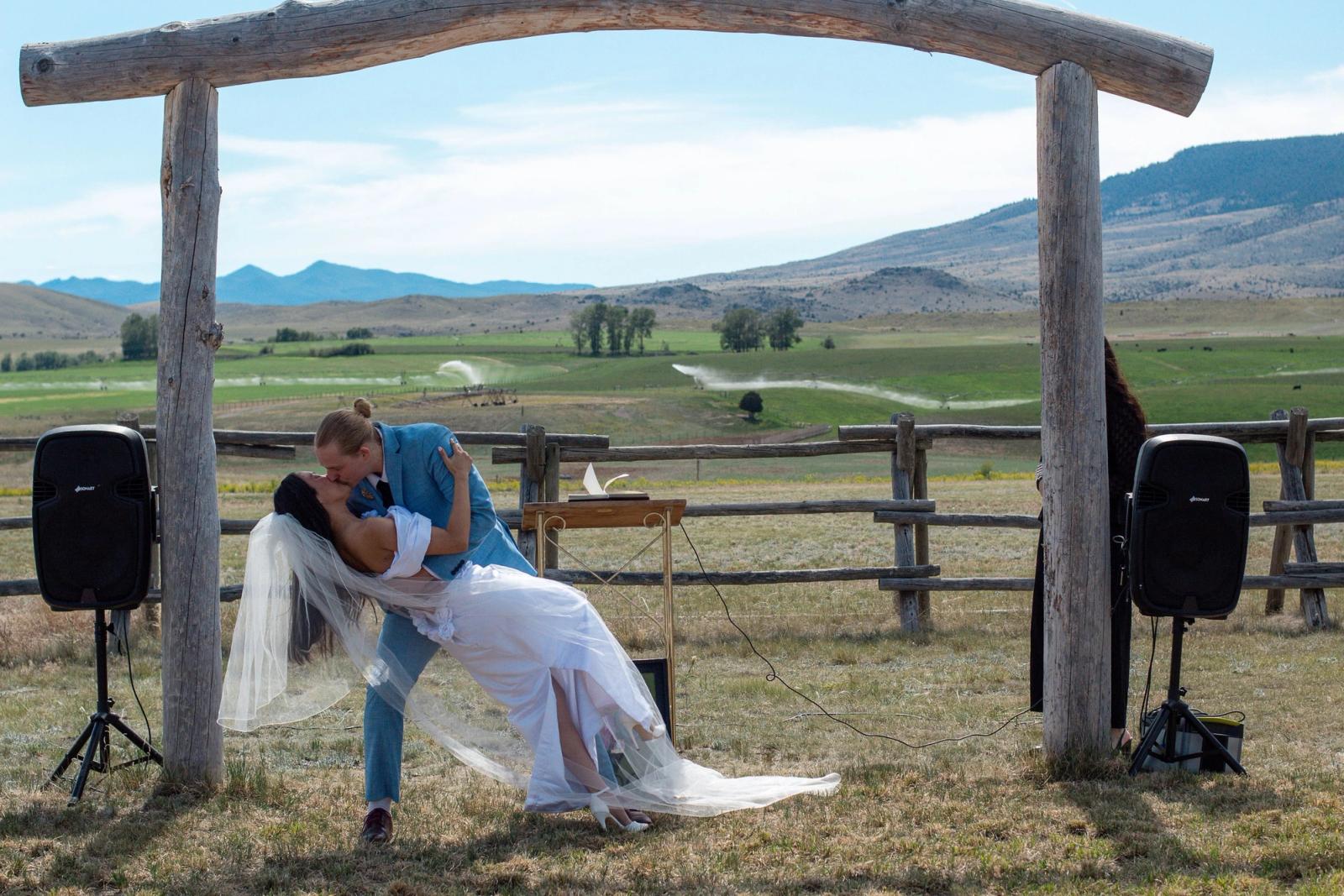 The first kiss: a groom dips his bride under the log arch as irrigation sprinklers arc across the valley