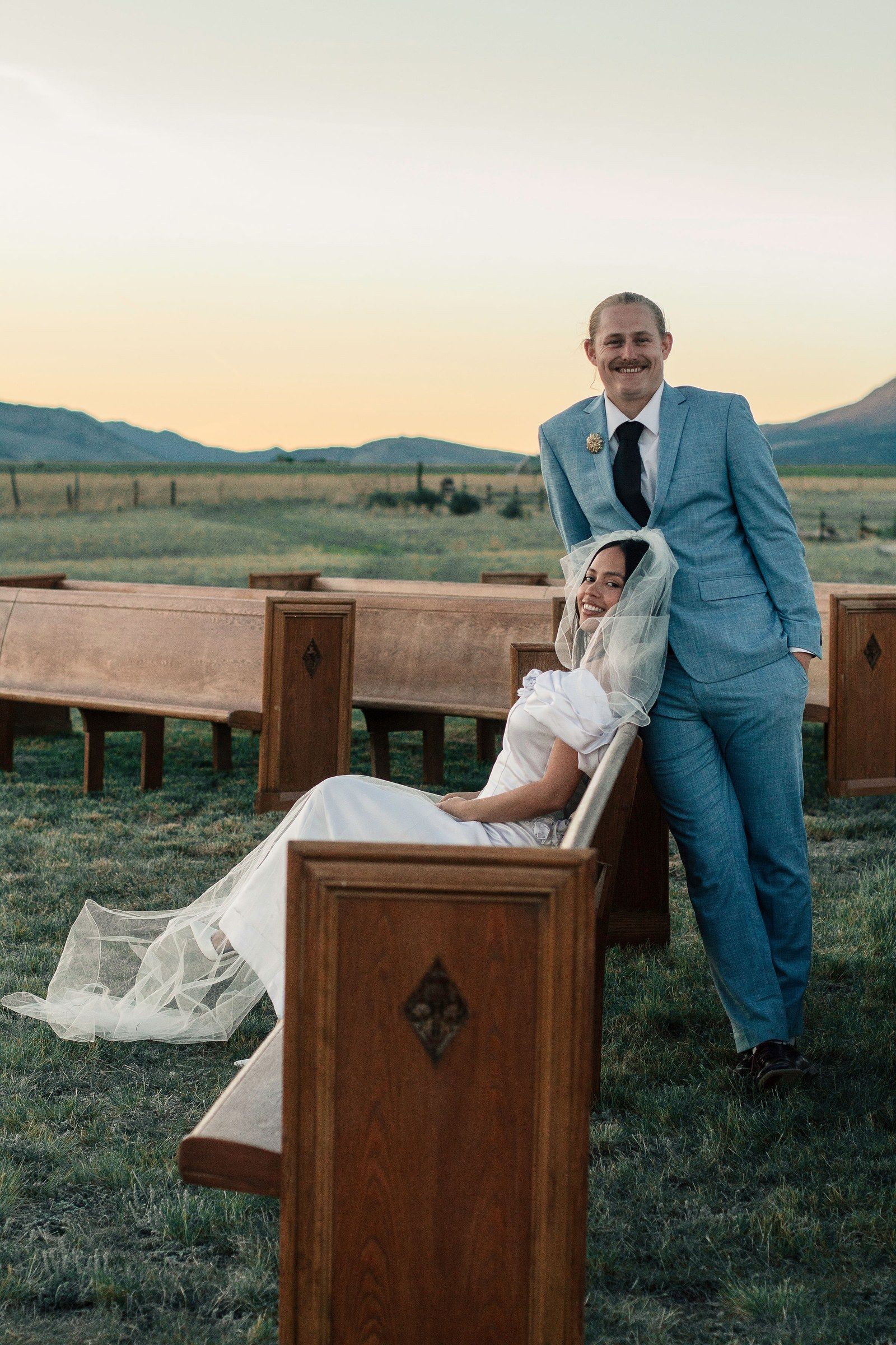 A groom stands by the meadow pews at sunset as his bride leans back laughing under her veil