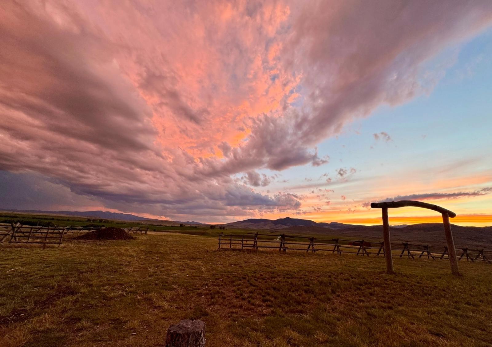 Pink and gold sunset clouds sweeping over the Ceremony Meadow, the log arch and jackleg fence against the evening sky