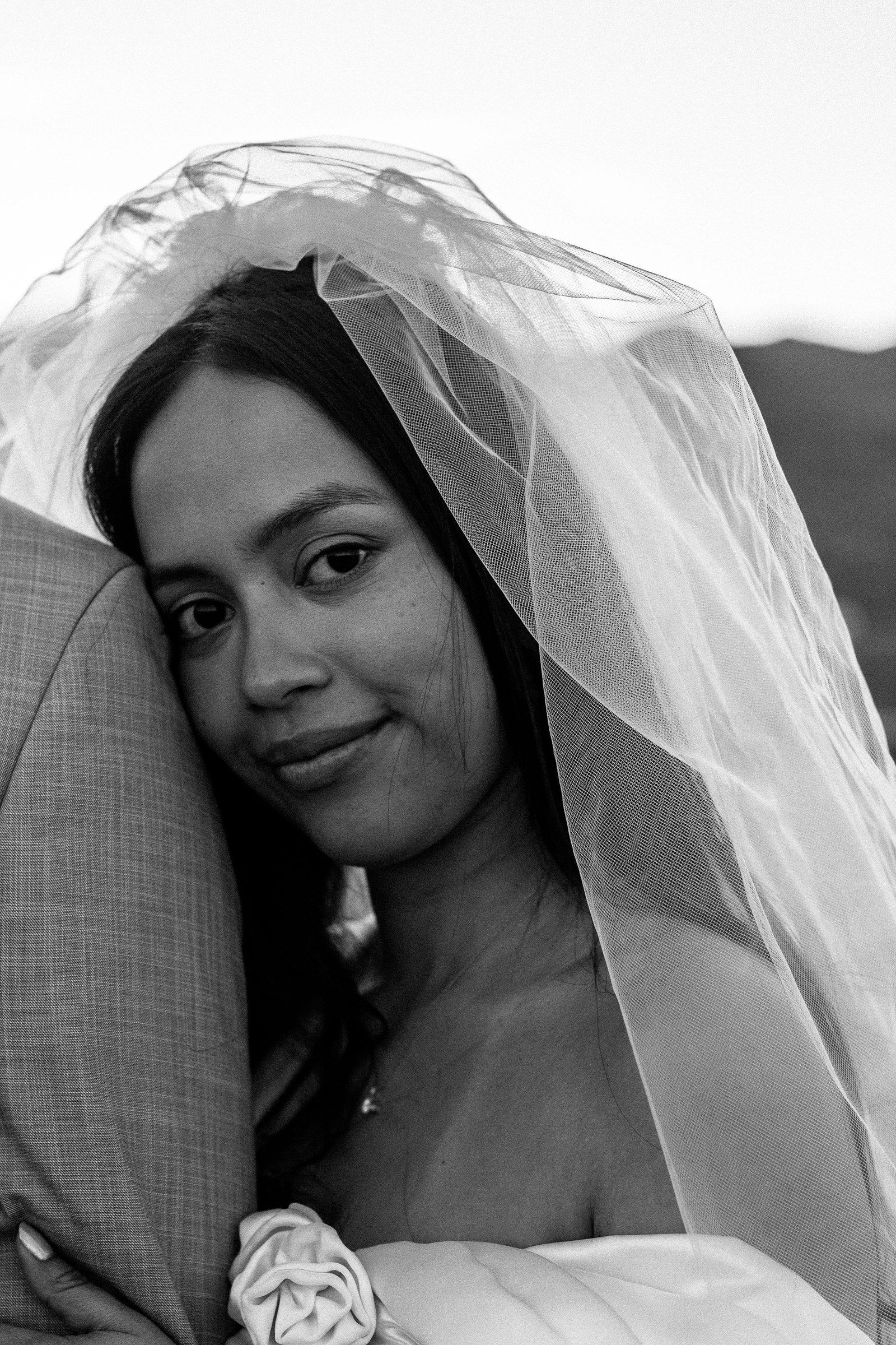 Black-and-white portrait of a bride under her veil, resting on her groom's shoulder