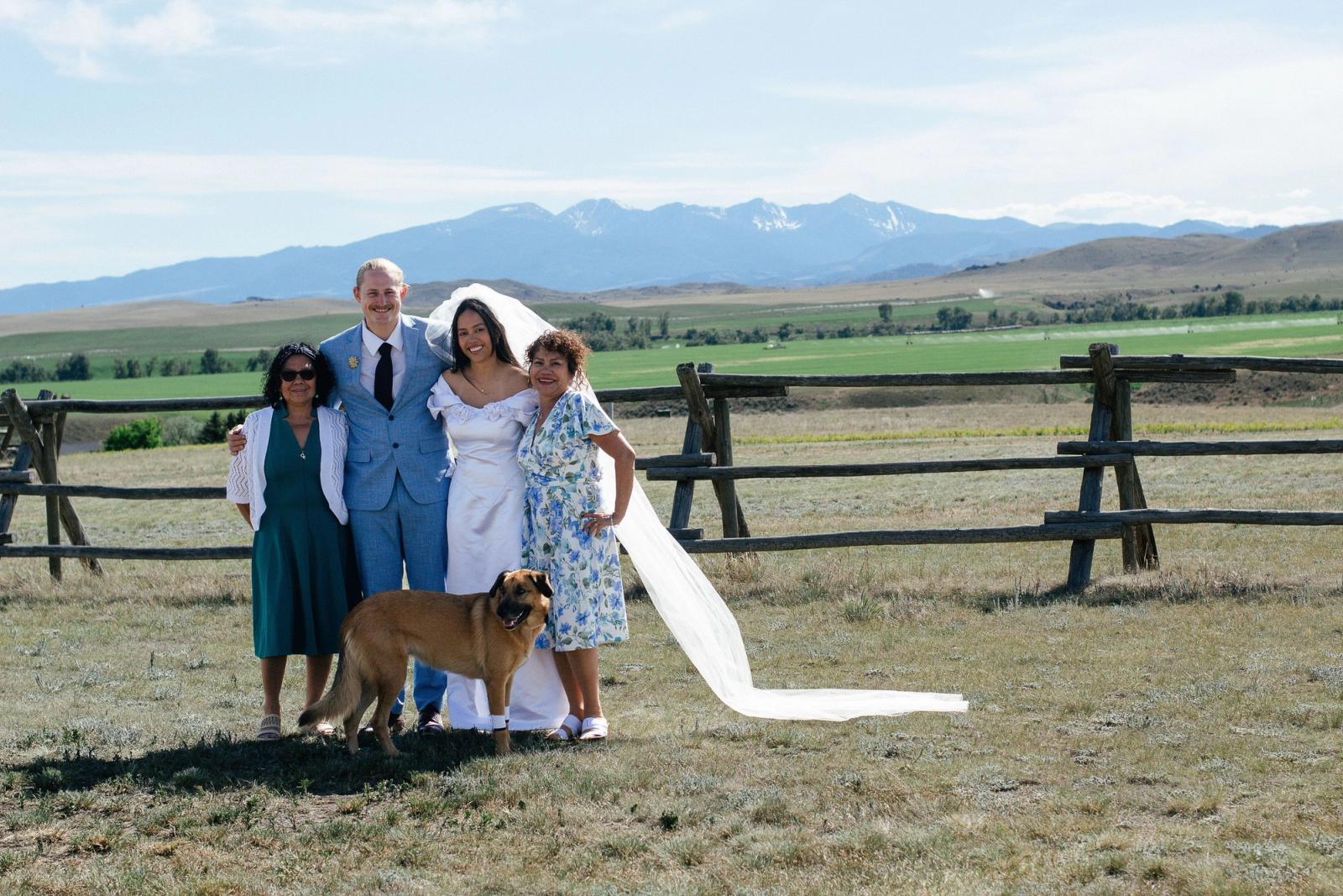 The newlyweds with family and a friendly dog at the fence, snow-dusted peaks beyond