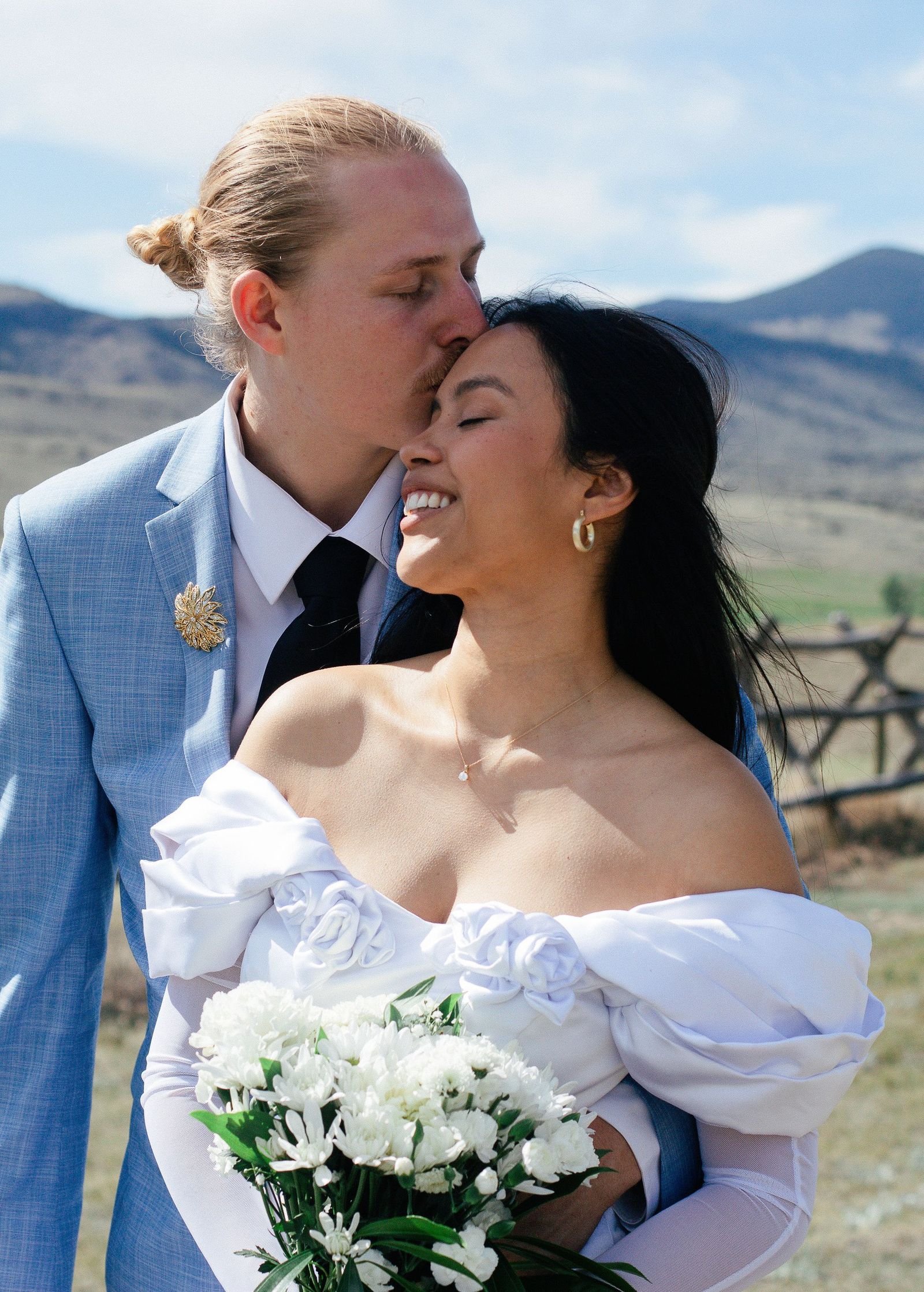 A groom kisses his bride's forehead, her white bouquet in hand and the mountains behind