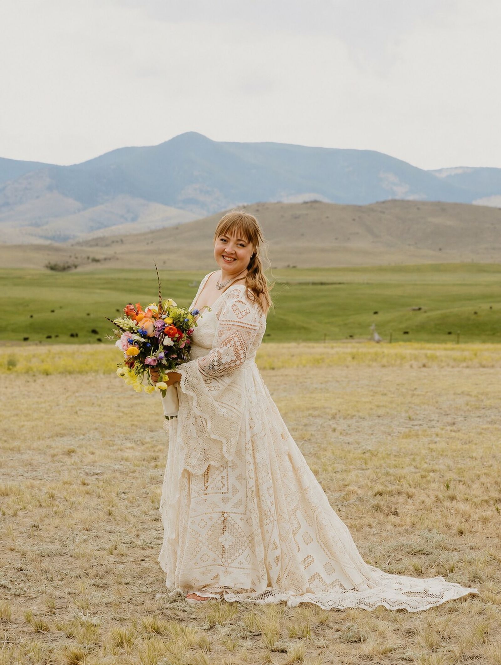 A bride in a lace gown holding a wildflower bouquet in the meadow, mountains behind