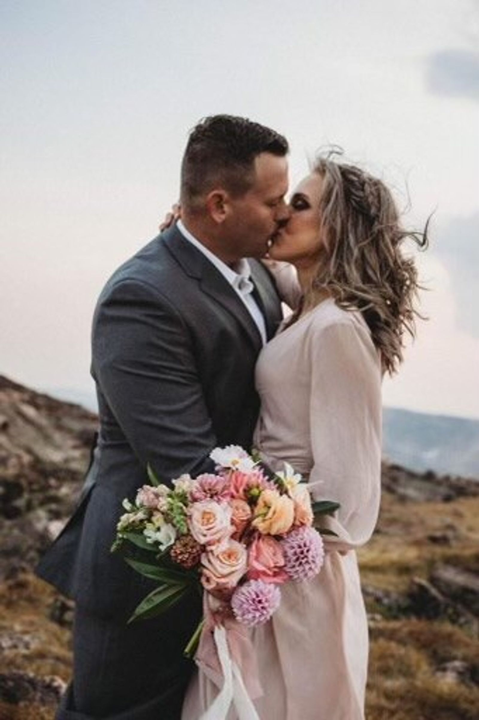 A couple kisses on a windswept ridge, her bouquet of peach roses and pink dahlias held between them