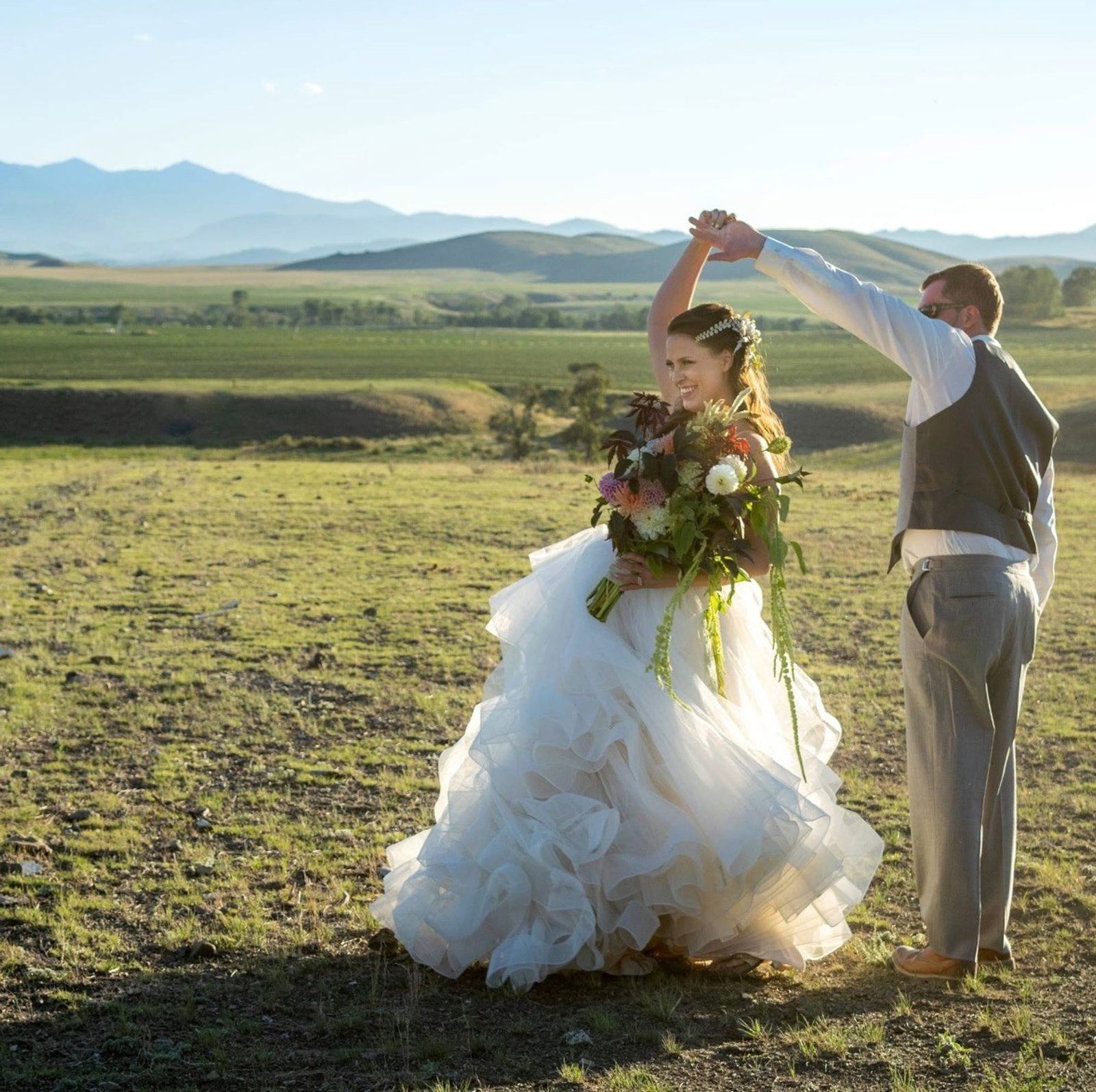 A groom twirls his bride in the meadow, the valley and mountains behind them