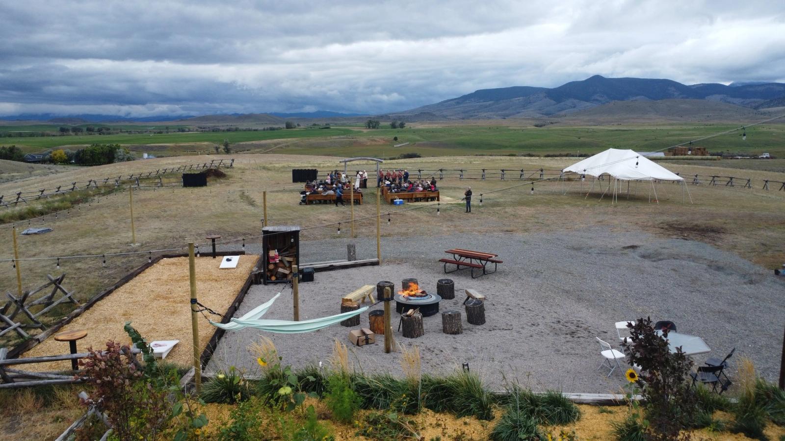 The fire pit and hammocks from above, with a ceremony gathered in the meadow and a white tent under the mountains