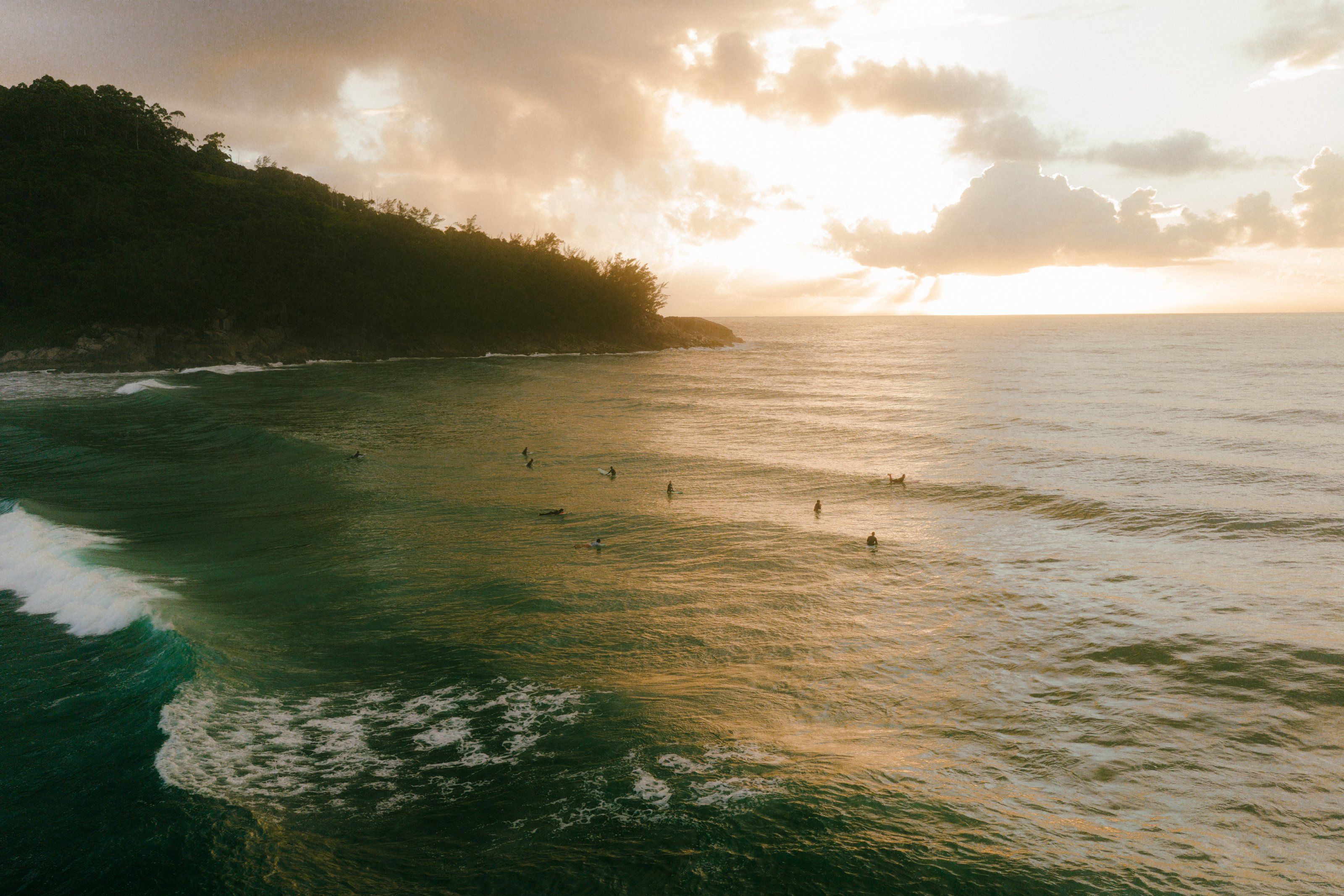 Vista aérea al atardecer de una playa del sur de Brasil, con surfistas esperando olas junto a un morro con vegetación