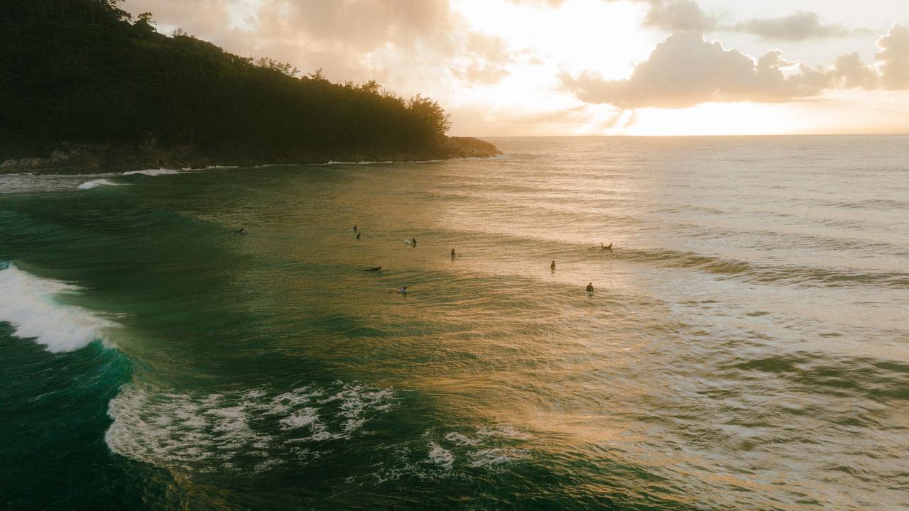 Vista aérea al atardecer de una playa del sur de Brasil, con surfistas esperando olas junto a un morro con vegetación