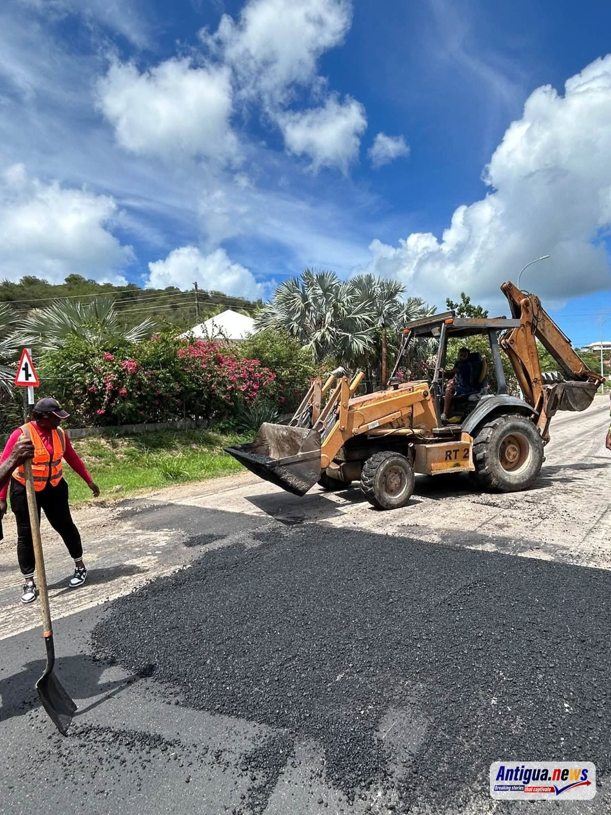 Road construction workers operate paving machinery while laying fresh asphalt along a roadway in a tropical Caribbean setting.